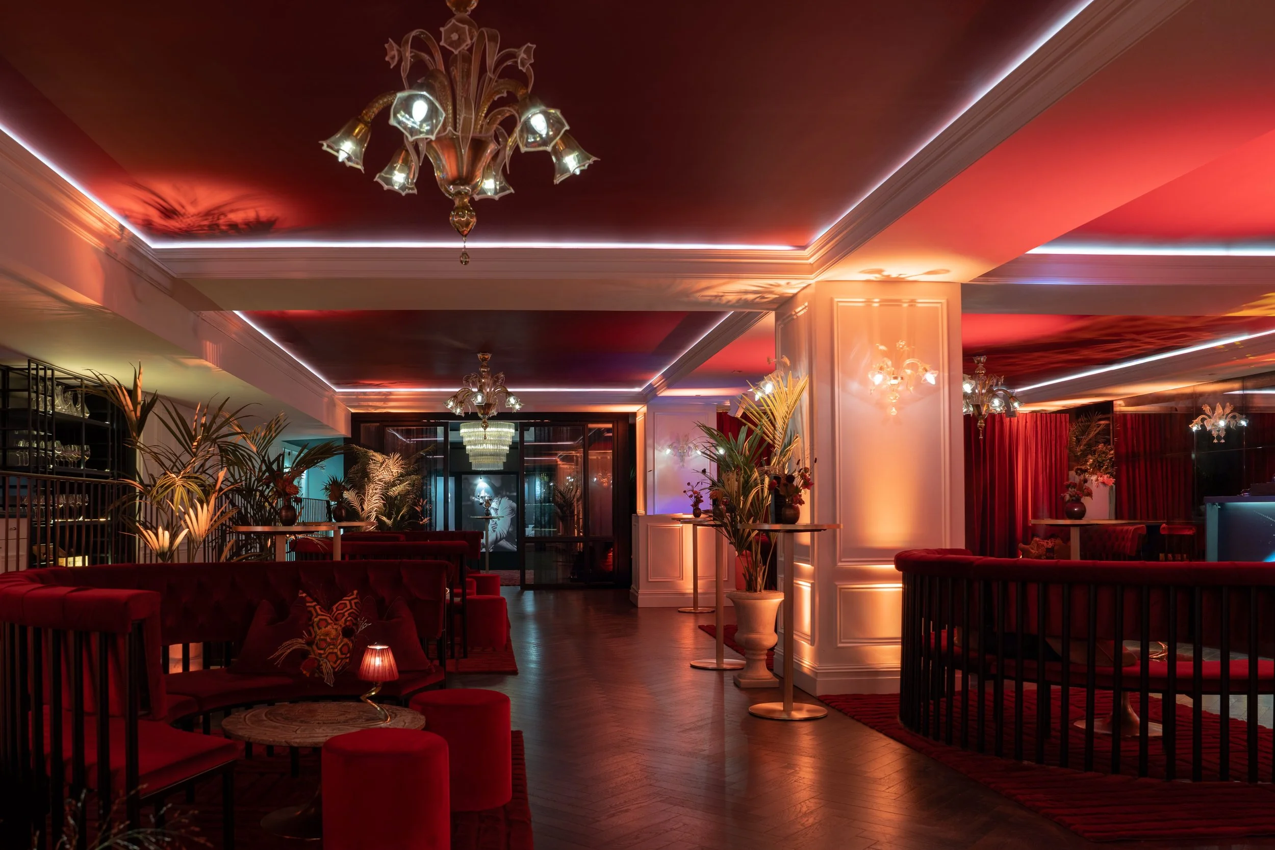Elegant lounge area with red velvet seating, potted plants, chandelier lighting, and decorative drapes, illuminated with warm, ambient lighting.