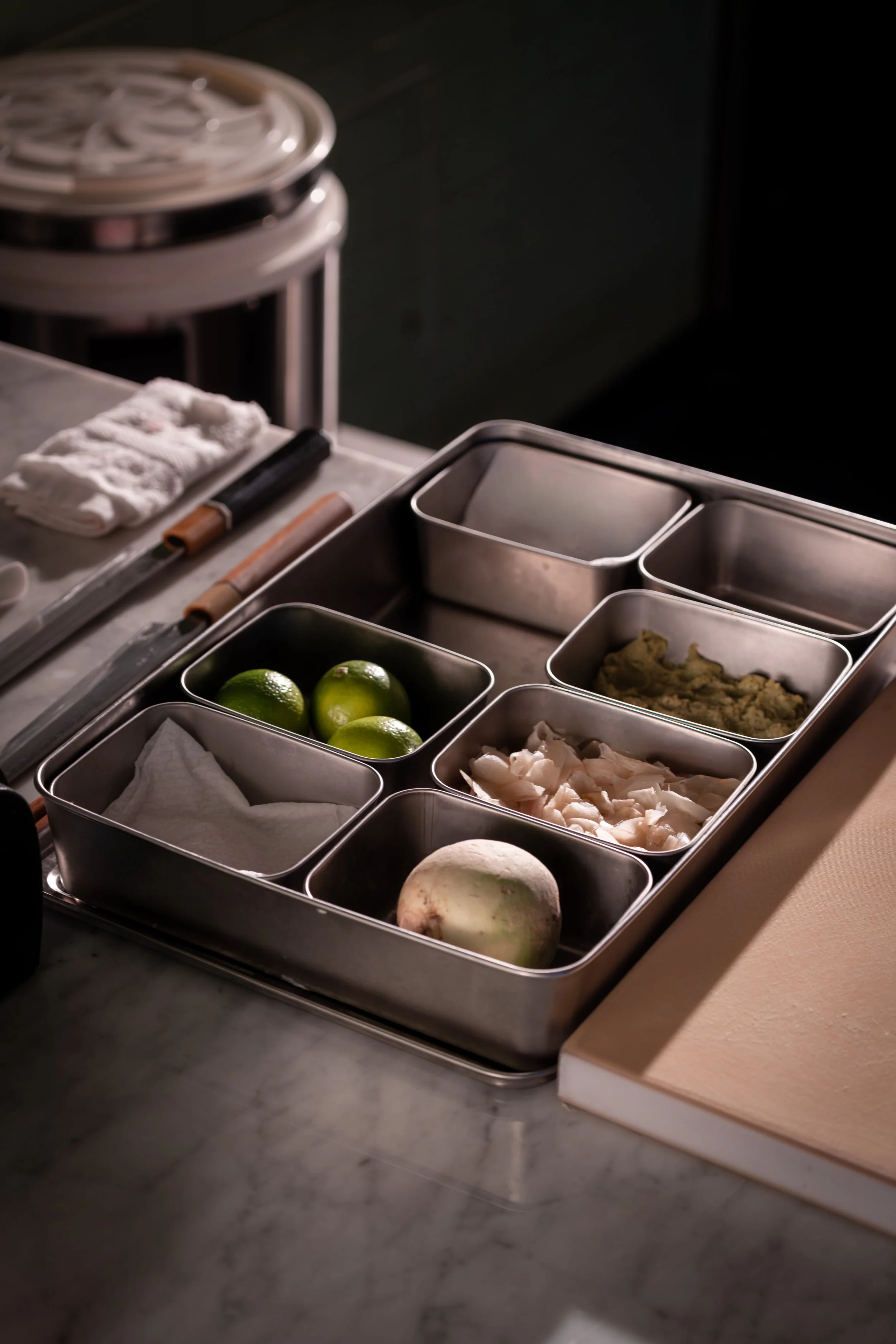 A metal tray with compartments holding limes, a turnip, chopped white onions, ginger paste, and a white cloth, on a marble countertop in a kitchen.