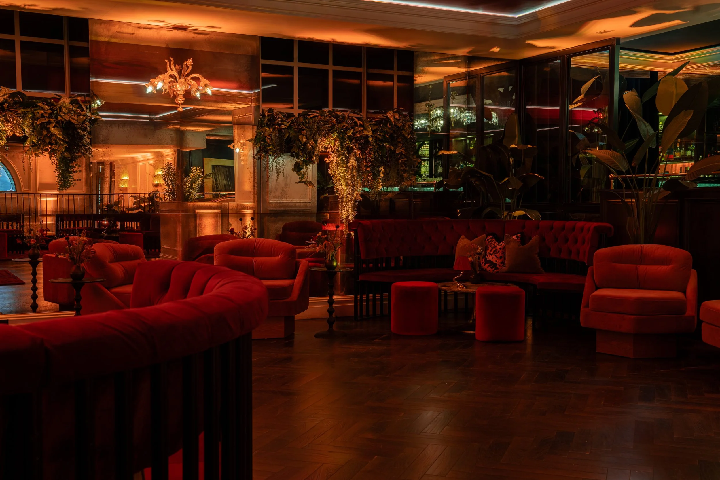 Interior of a dimly lit lounge with plush red velvet chairs and sofas, decorative plants, a chandelier, and warm lighting reflections on glass walls.