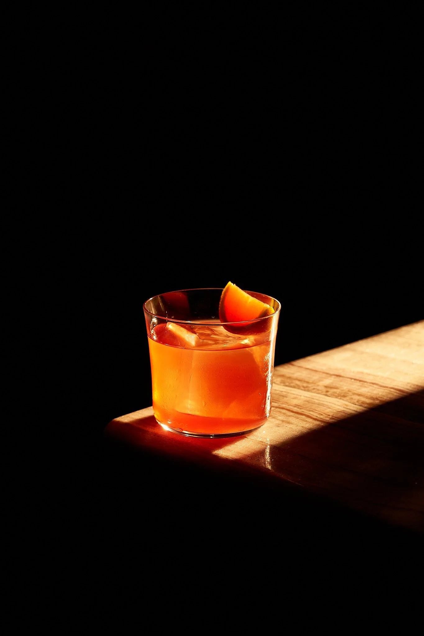 A glass of orange-colored cocktail with a slice of orange on the rim, placed on a wooden surface against a dark background.