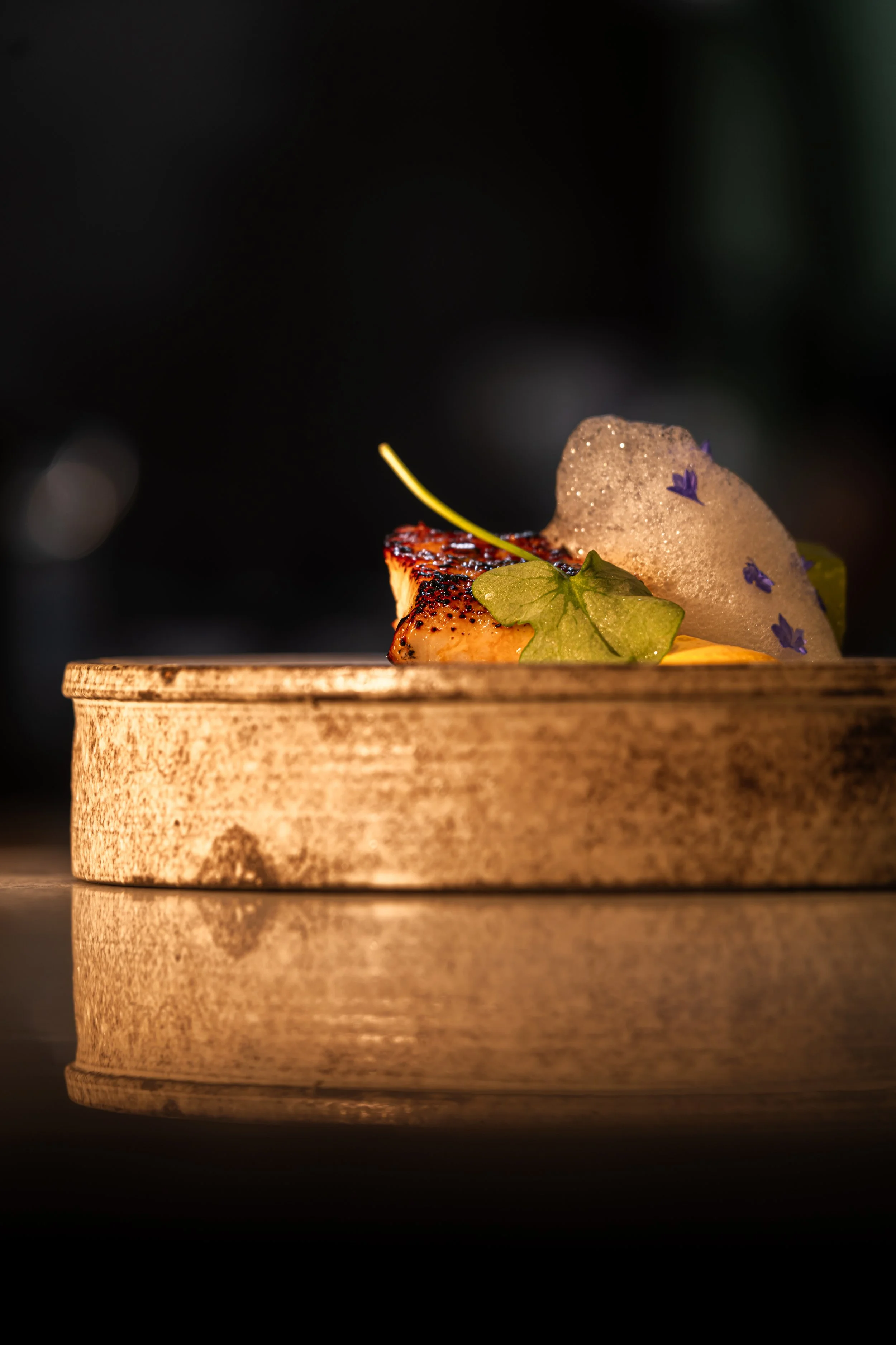 A close-up of a gourmet dish served in a round wooden container, featuring a piece of seared fish, topped with foam, green garnish, and edible purple flowers, with a dark background and reflective surface.