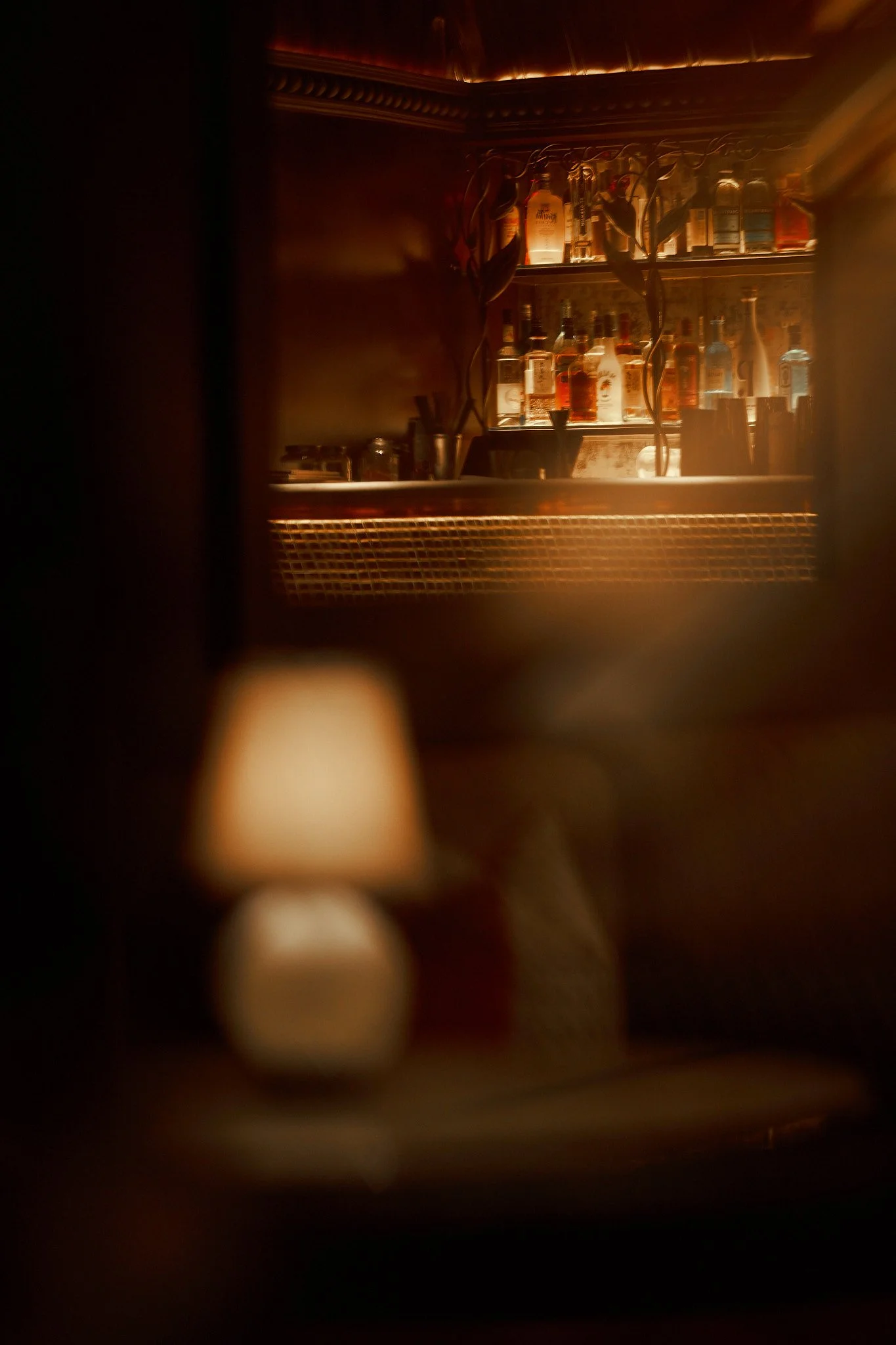 A blurry, dimly-lit interior scene with a small table lamp and a blurred bar shelf with bottles in the background.