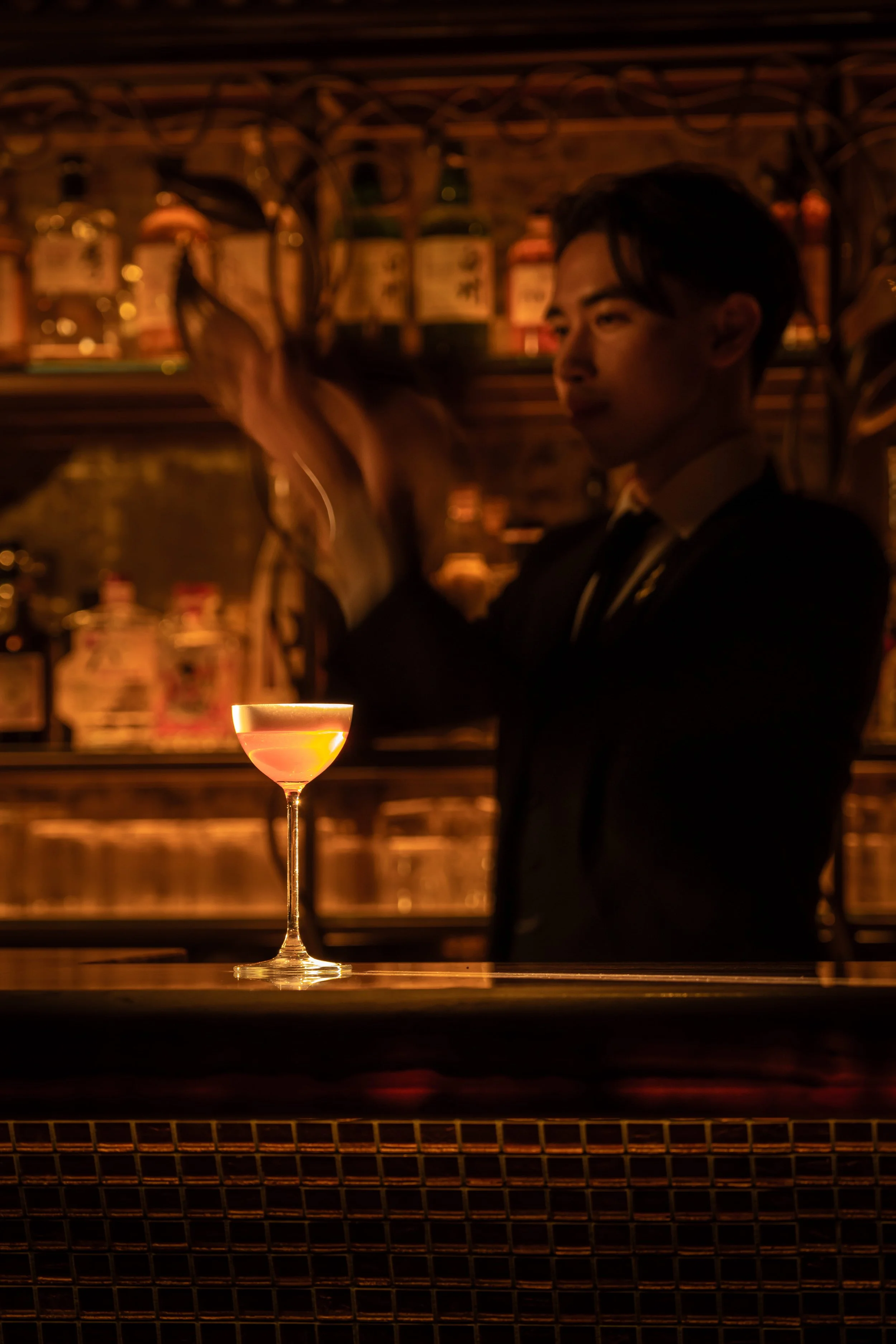 A bartender in a dark suit prepares a cocktail behind a bar, with a coupe glass filled with a pinkish-orange drink on the counter in front. Shelves with bottles are visible in the background.
