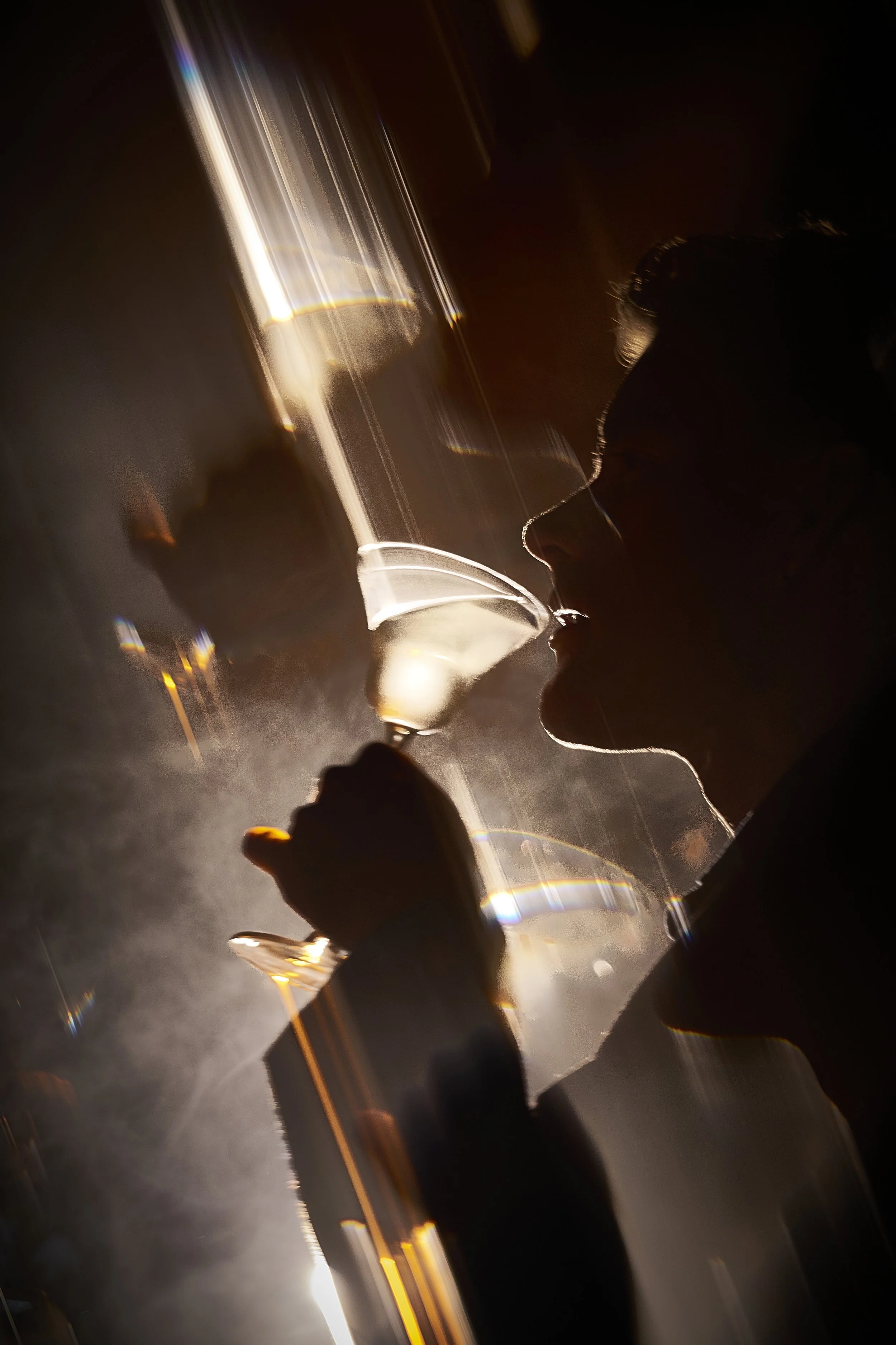 Person drinking from a wine glass in a dimly lit setting, silhouetted with streaks of light creating a dramatic effect.
