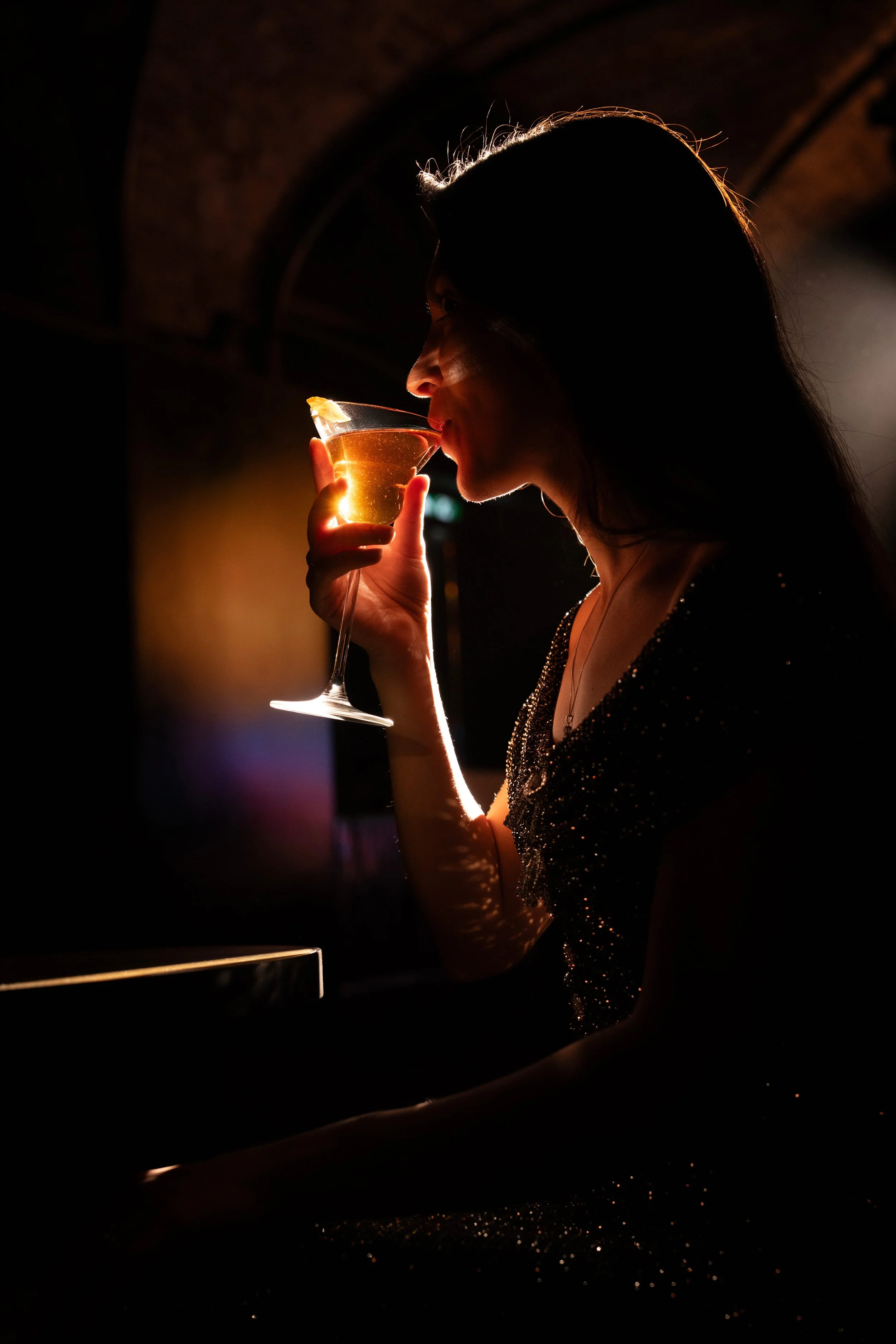 A woman with long hair and a sparkly dress enjoying a drink in a dark, stylish setting, illuminated by a warm backlight.