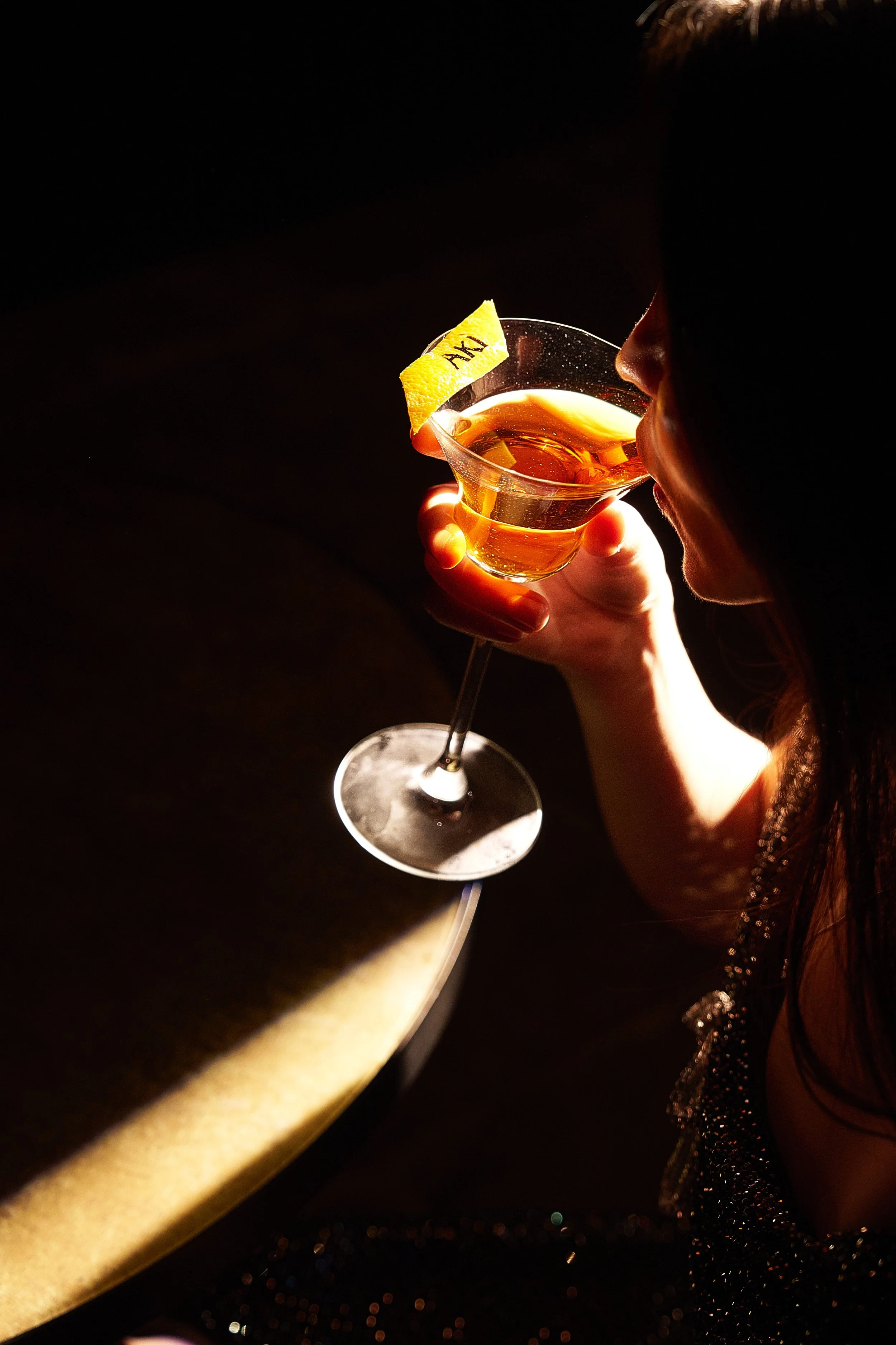 A woman with dark hair, wearing a sparkly dress, is holding a glass of amber-colored cocktail garnished with a lemon peel with the word "AM" on it, near her lips in a dimly lit setting.