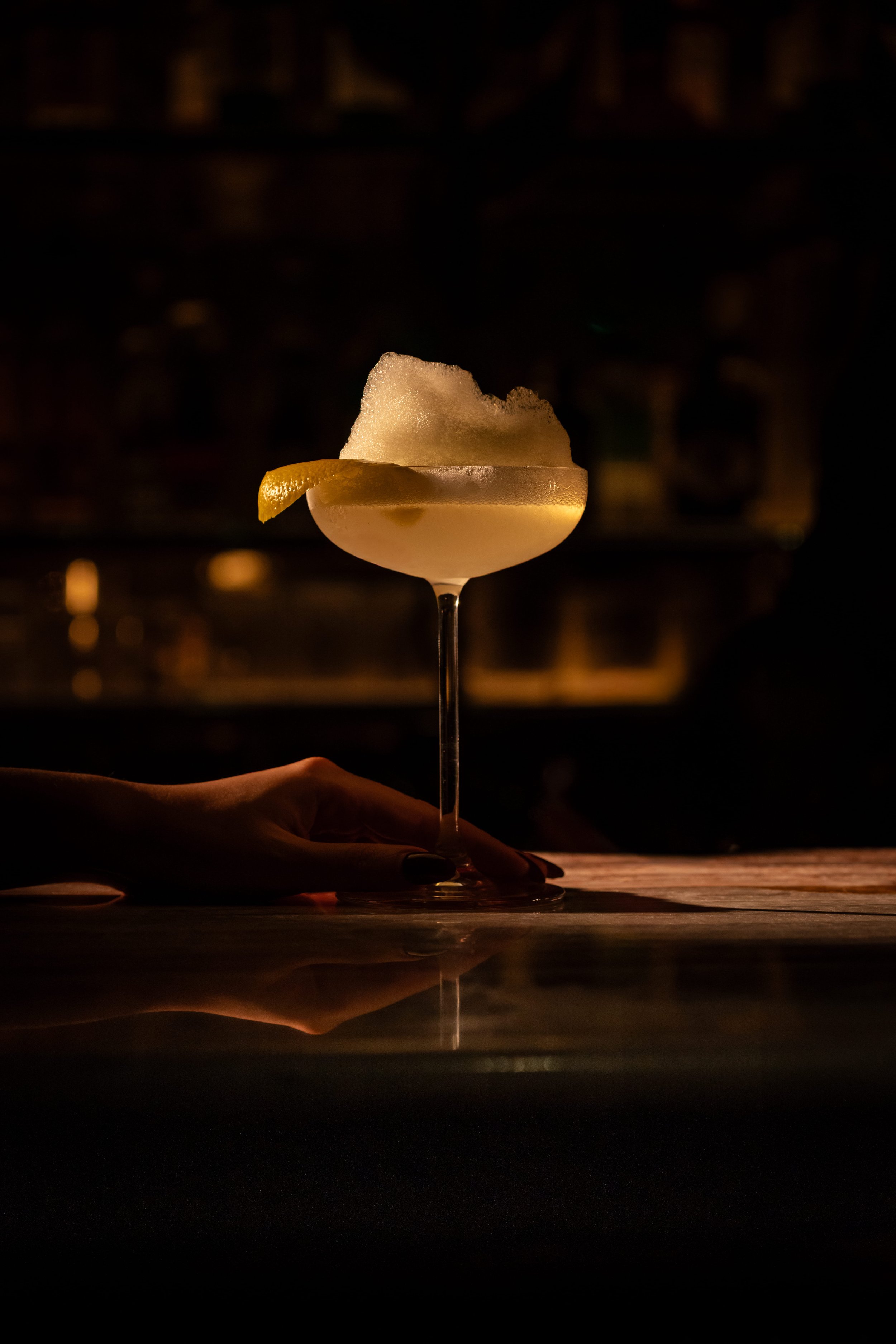 A cocktail glass with a lemon wedge garnish, filled with a foamy drink, being held by a person's hand in a dimly lit bar.
