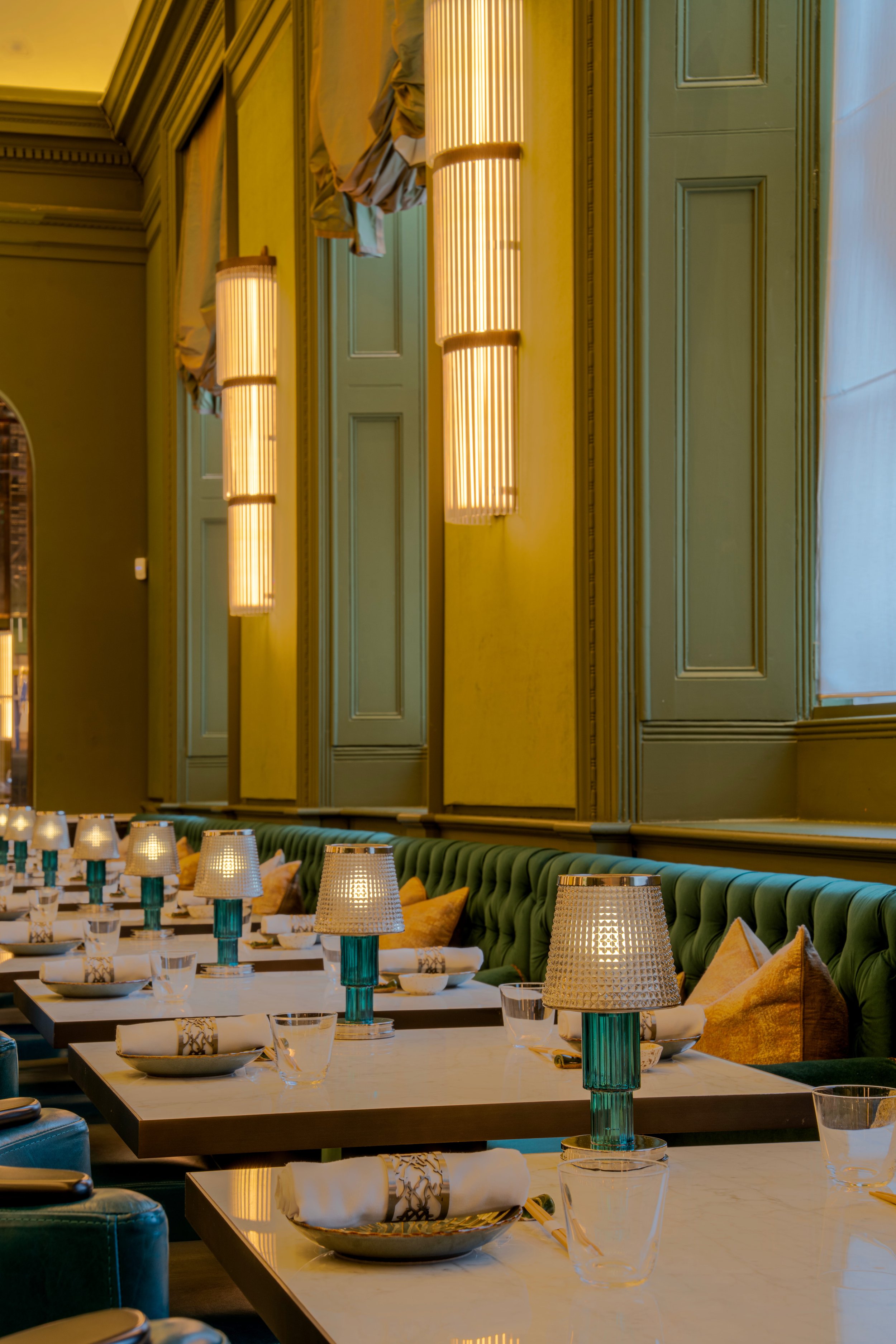 Elegant restaurant dining area with marble tables, green upholstered seating, and modern table lamps with textured shades and turquoise bases.