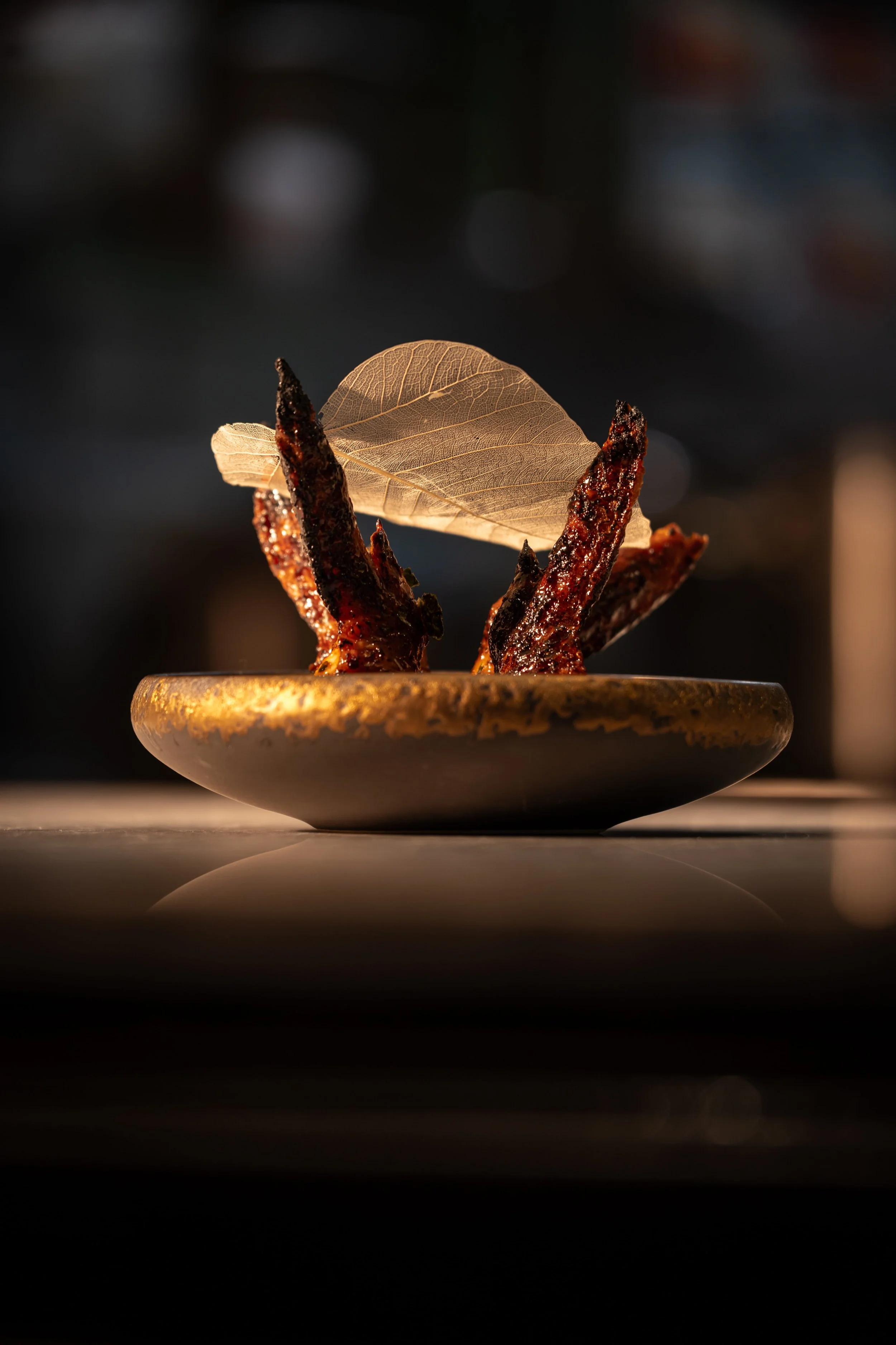 Close-up of a small dish with cooked chicken wings, garnished with a large leaf and a decorative leaf on top, with a warm, soft light highlighting the dish.