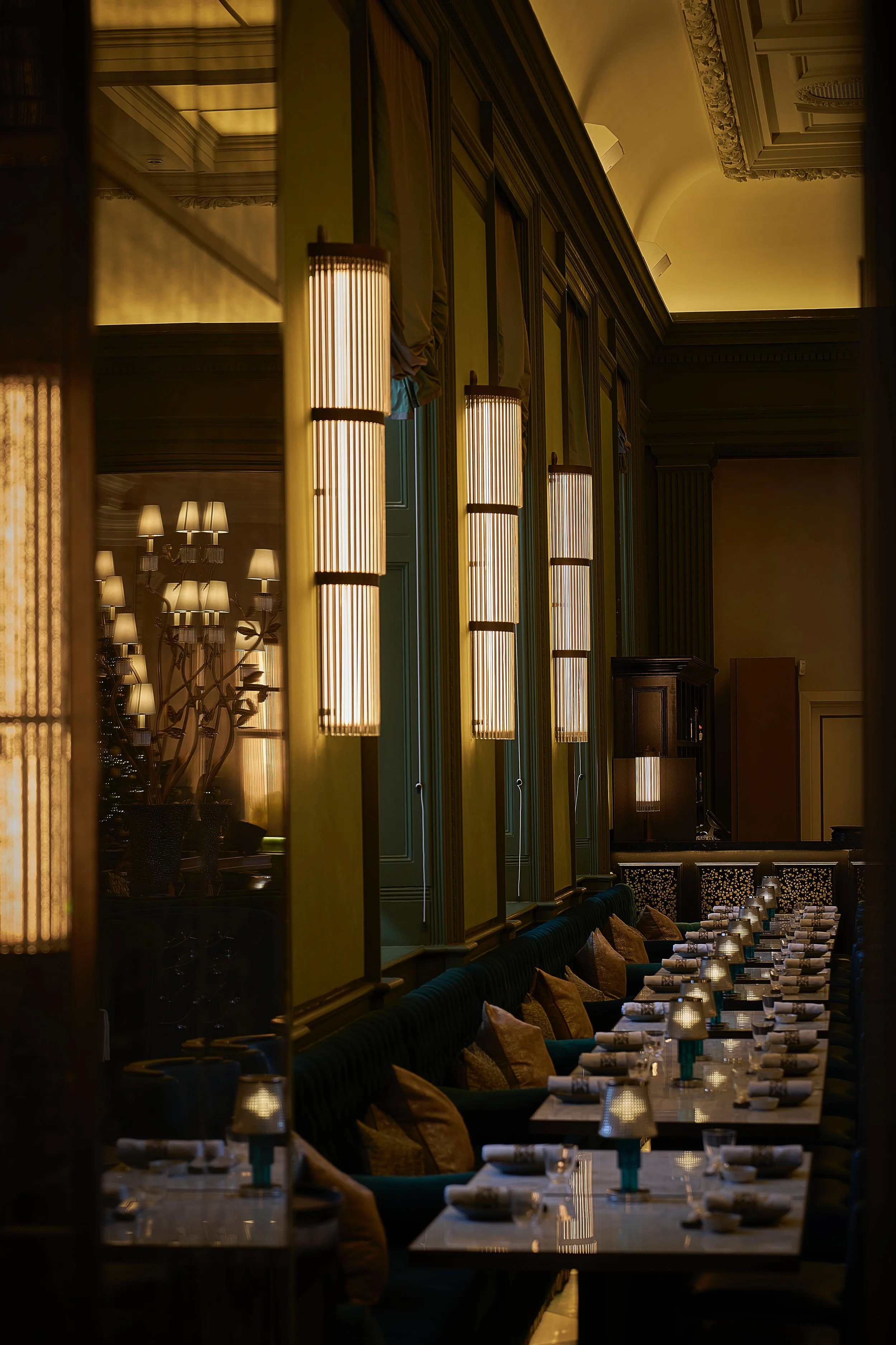 Elegant restaurant interior with a row of lit wall sconces, tables set with napkins and glasses, and plush upholstered seating with pillows.