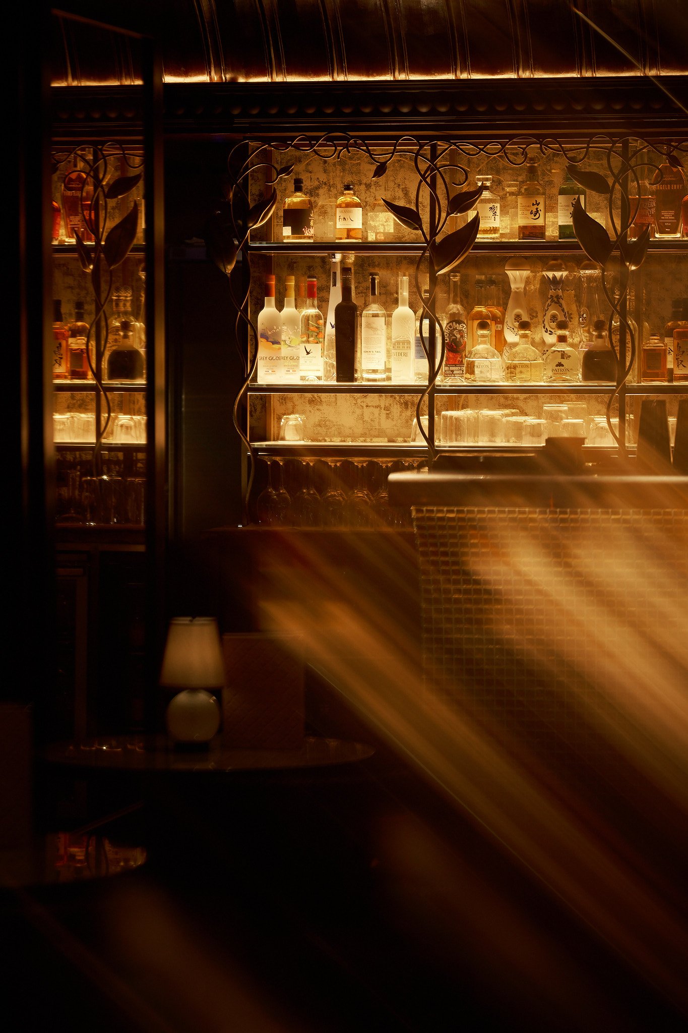 A bar shelf with various bottles of alcohol, illuminated with warm lighting, and decorative wrought iron leaves in front of the shelf.