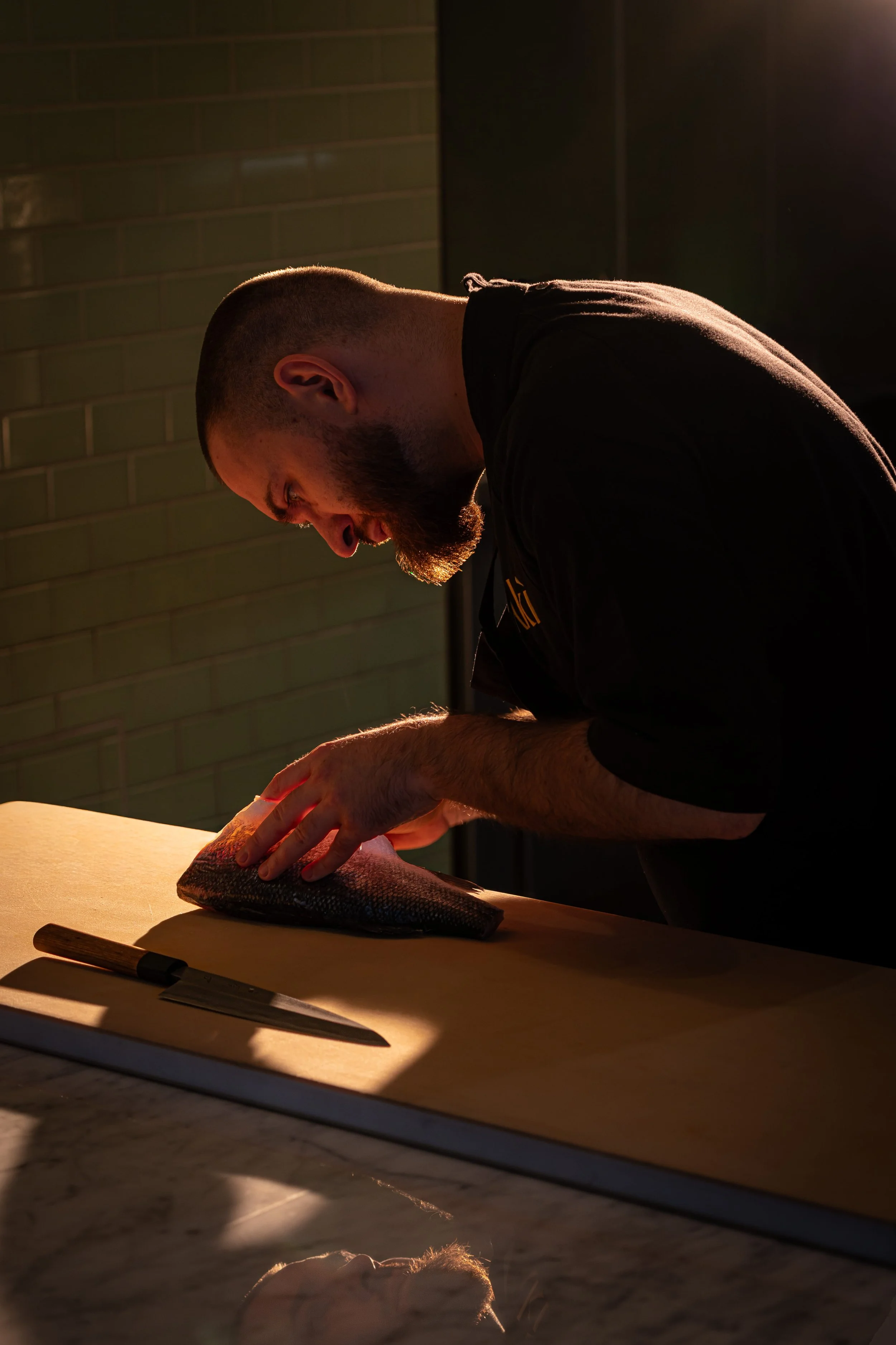 A man preparing a fish on a wooden cutting board, with a chef's knife nearby, in a dimly lit kitchen.