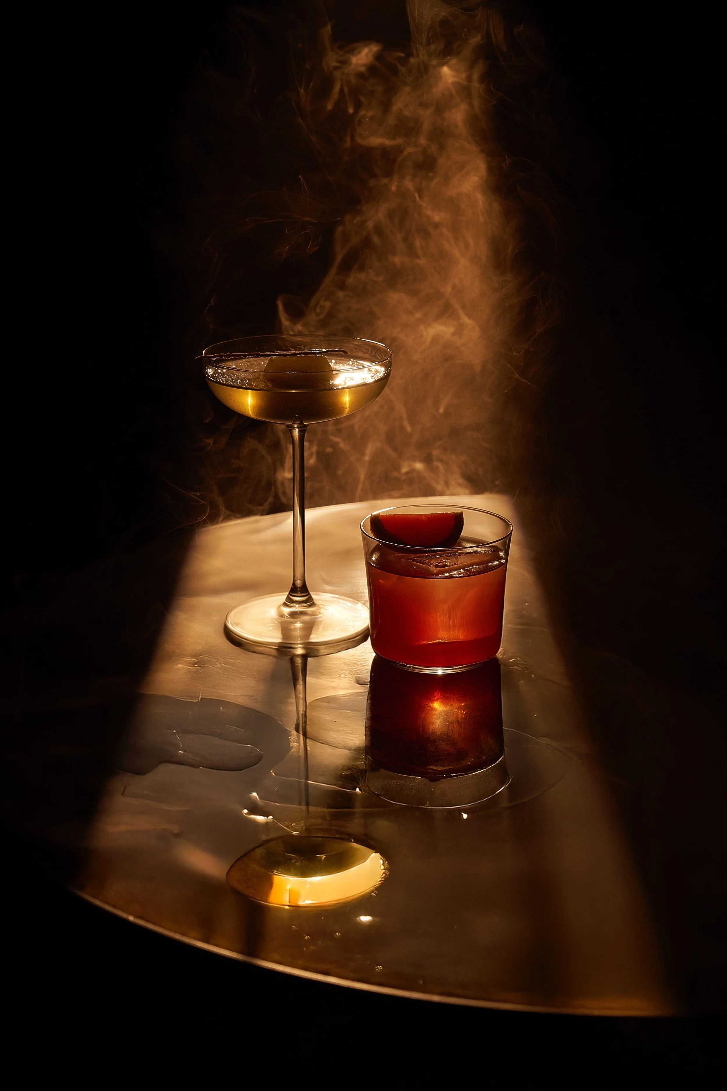 A martini cocktail in a coupe glass and a whiskey on the rocks in a short glass with a lime wedge, placed on a reflective metallic surface with a smoky background.