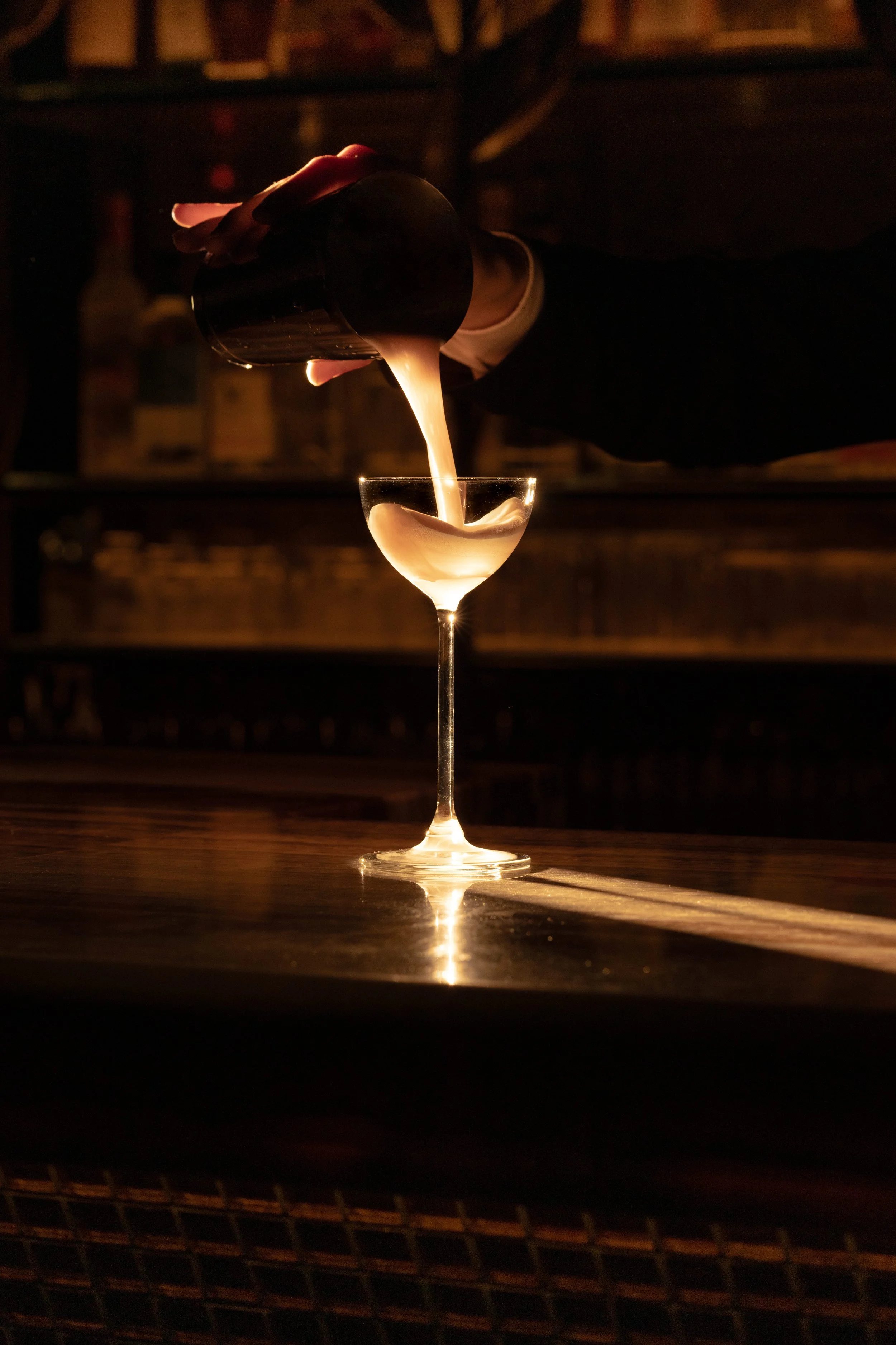 A bartender pours a creamy cocktail from a shaker into a martini glass at a dimly lit bar.