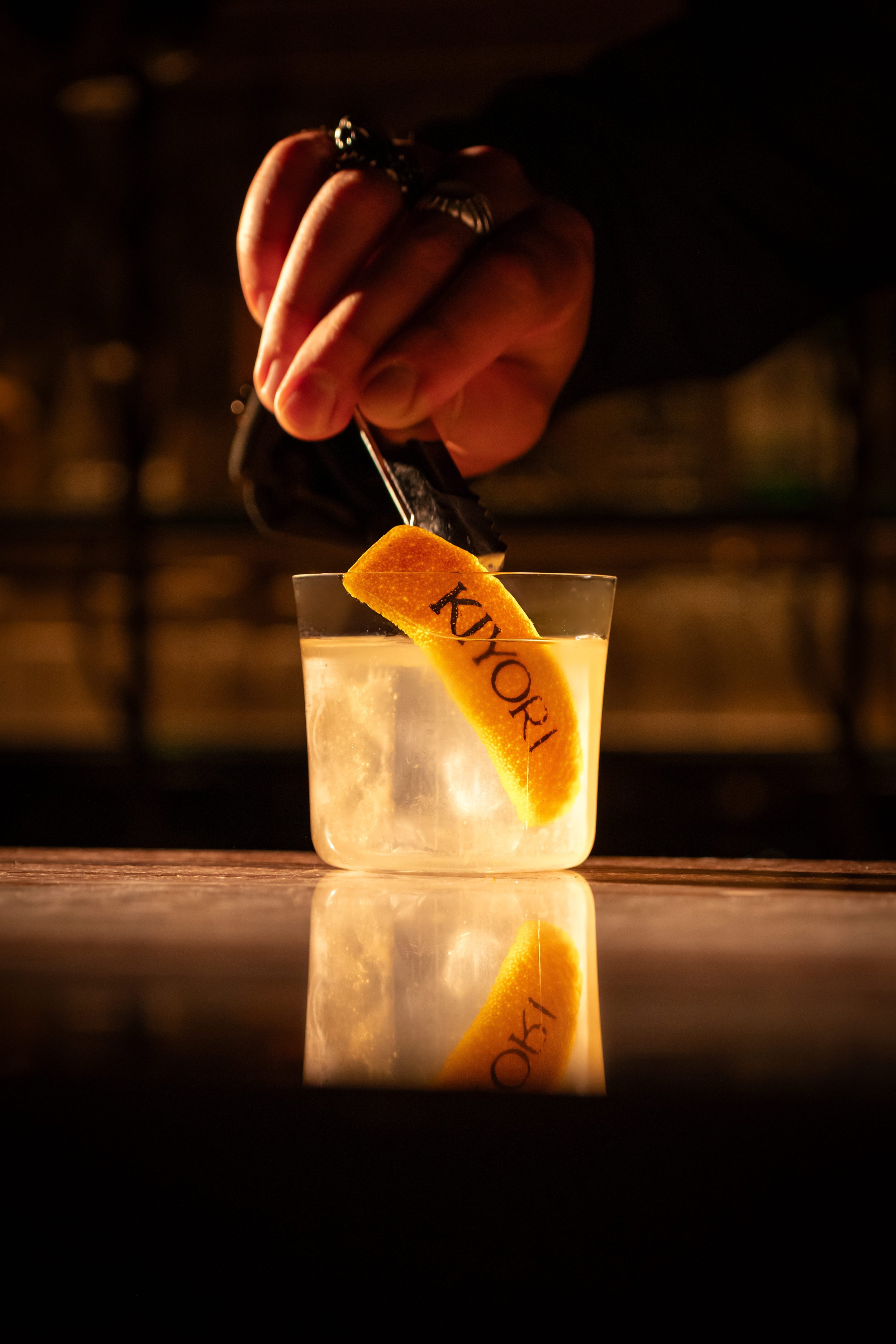 A hand with rings is placing a black object into a glass of clear liquid, which has an orange slice with the word 'KYOTO' written on it, reflecting on a table.