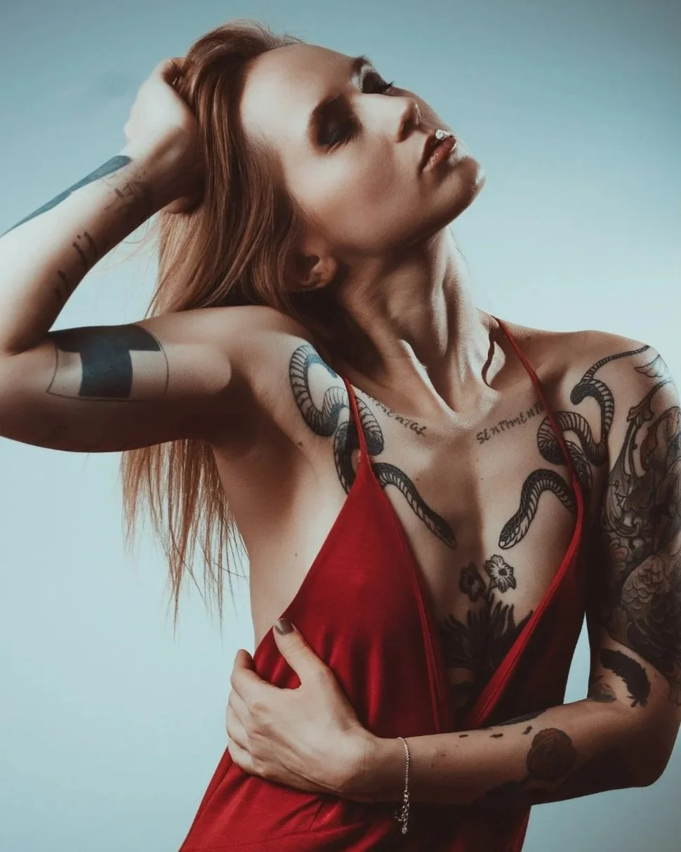 Eine Frau mit roten Haaren trägt ein rotes Kleid und hat viele Tätowierungen auf Armen, Schulter, Brust und Bauch. Sie ist in eleganter Pose mit geschlossenen Augen, die Hand hinter dem Kopf und der anderen Hand auf der Brust.