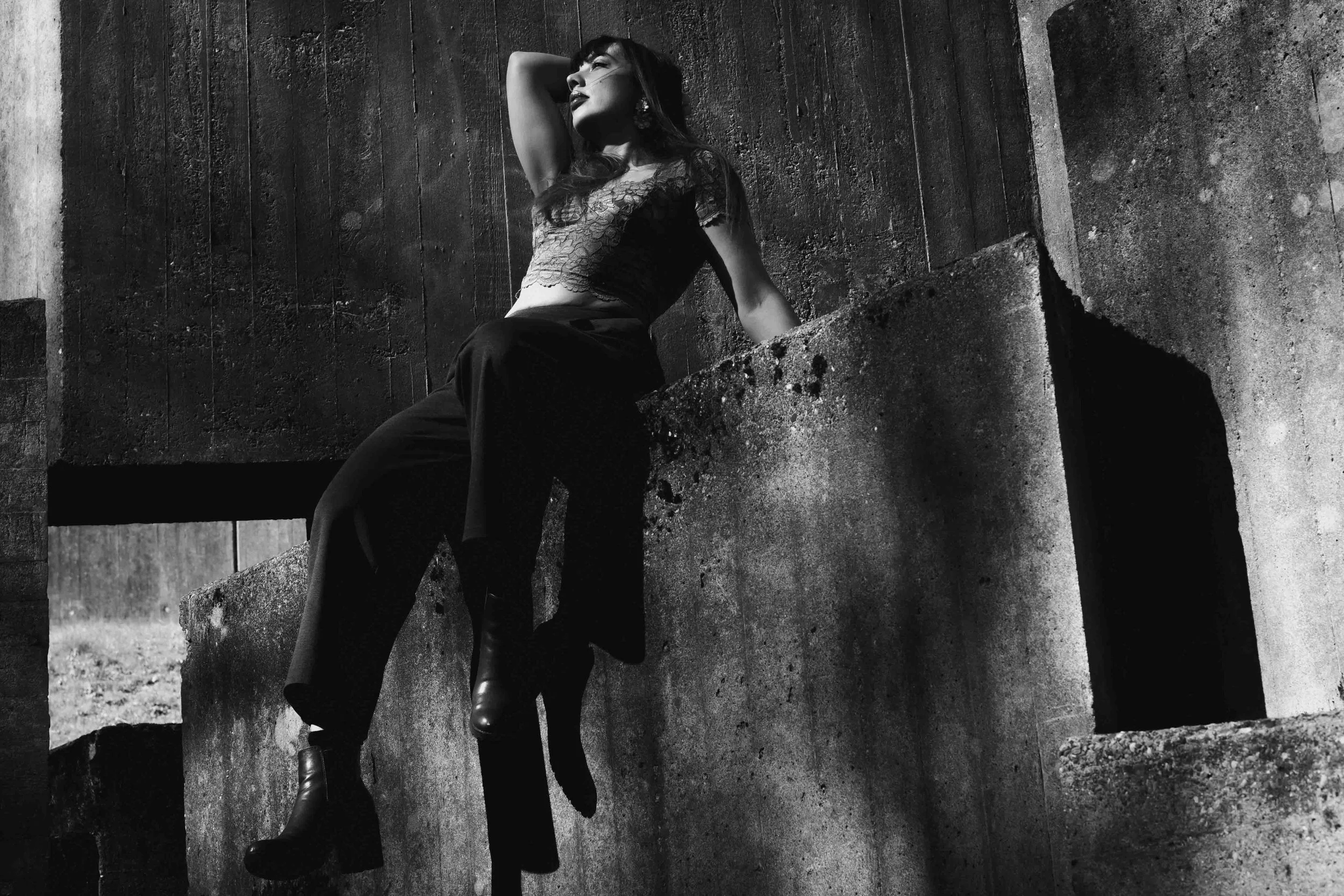 Schwarz-weiß-Fotografie einer Frau, die auf einer verfallenen Betonmauer lehnt, mit einem grauen Holzhintergrund im Freien, die Pose ist entspannt und nachdenklich.