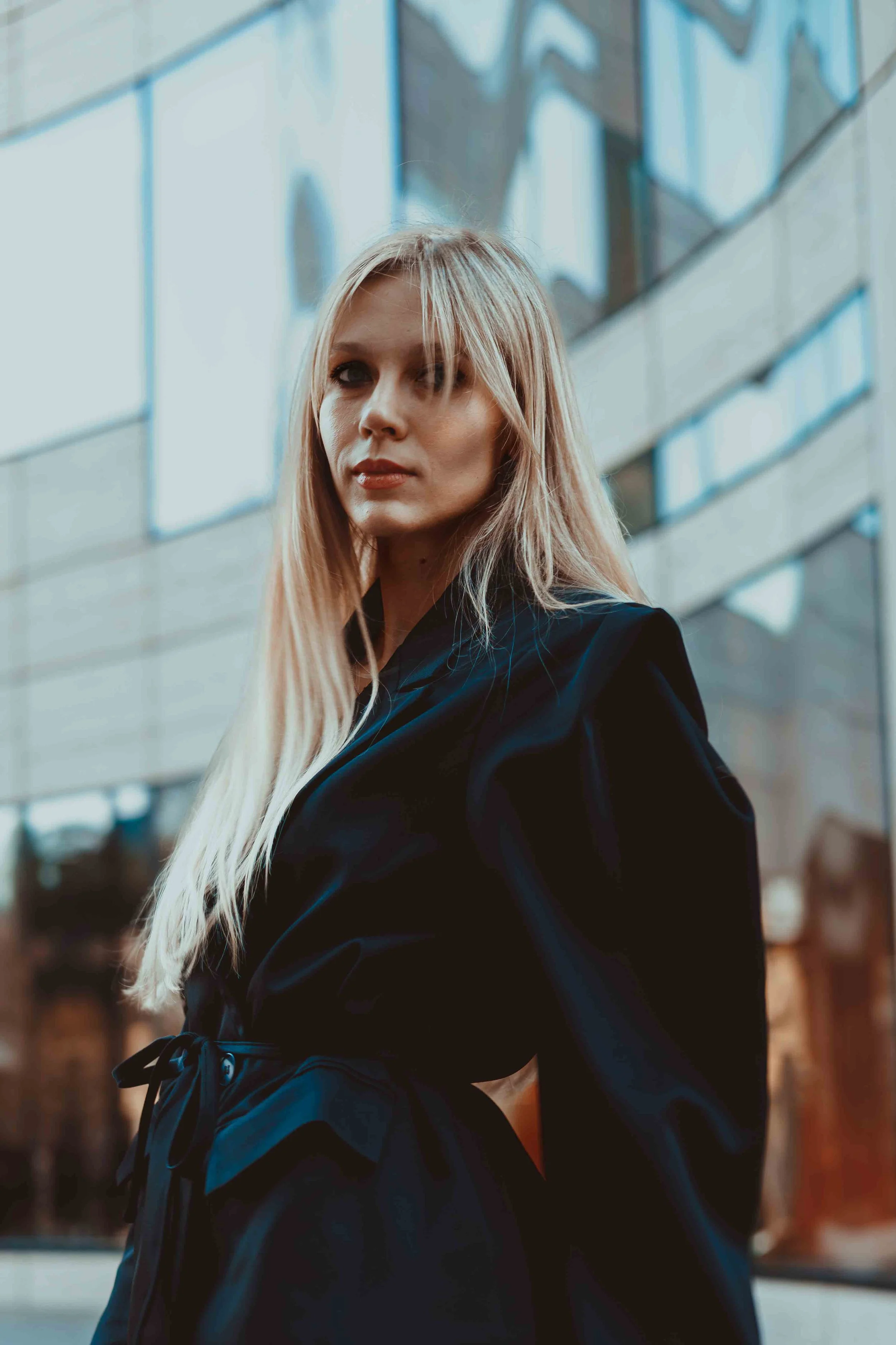 Eine junge Frau mit langem blondem Haar steht vor einem modernen Gebäude mit großen Glasscheiben. Sie trägt einen schwarzen Mantel und blickt ernst in die Kamera.