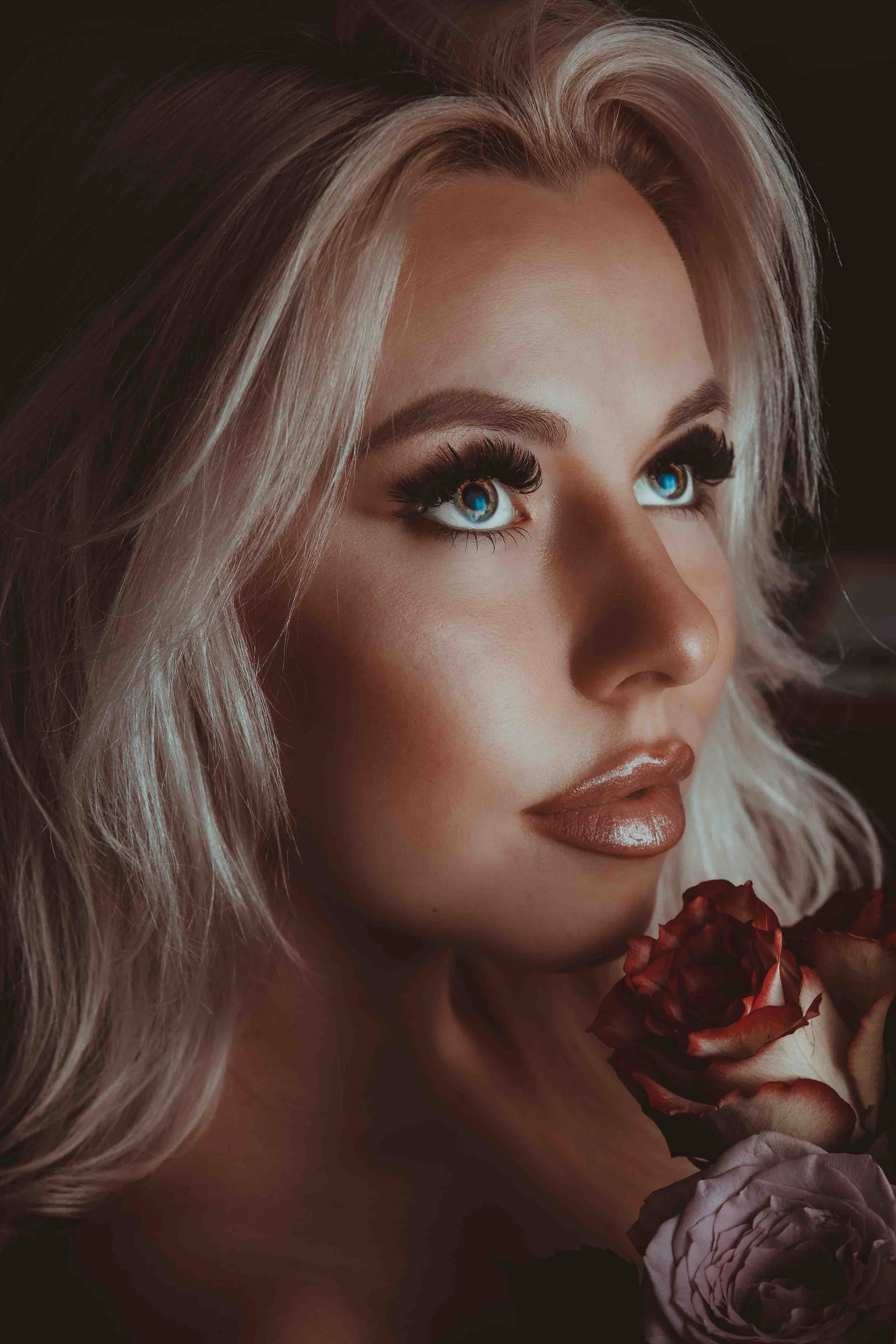Nahaufnahme eines Mädchens mit blonden, lockigen Haaren, blauen Augen und Make-up, das eine Rose hält.