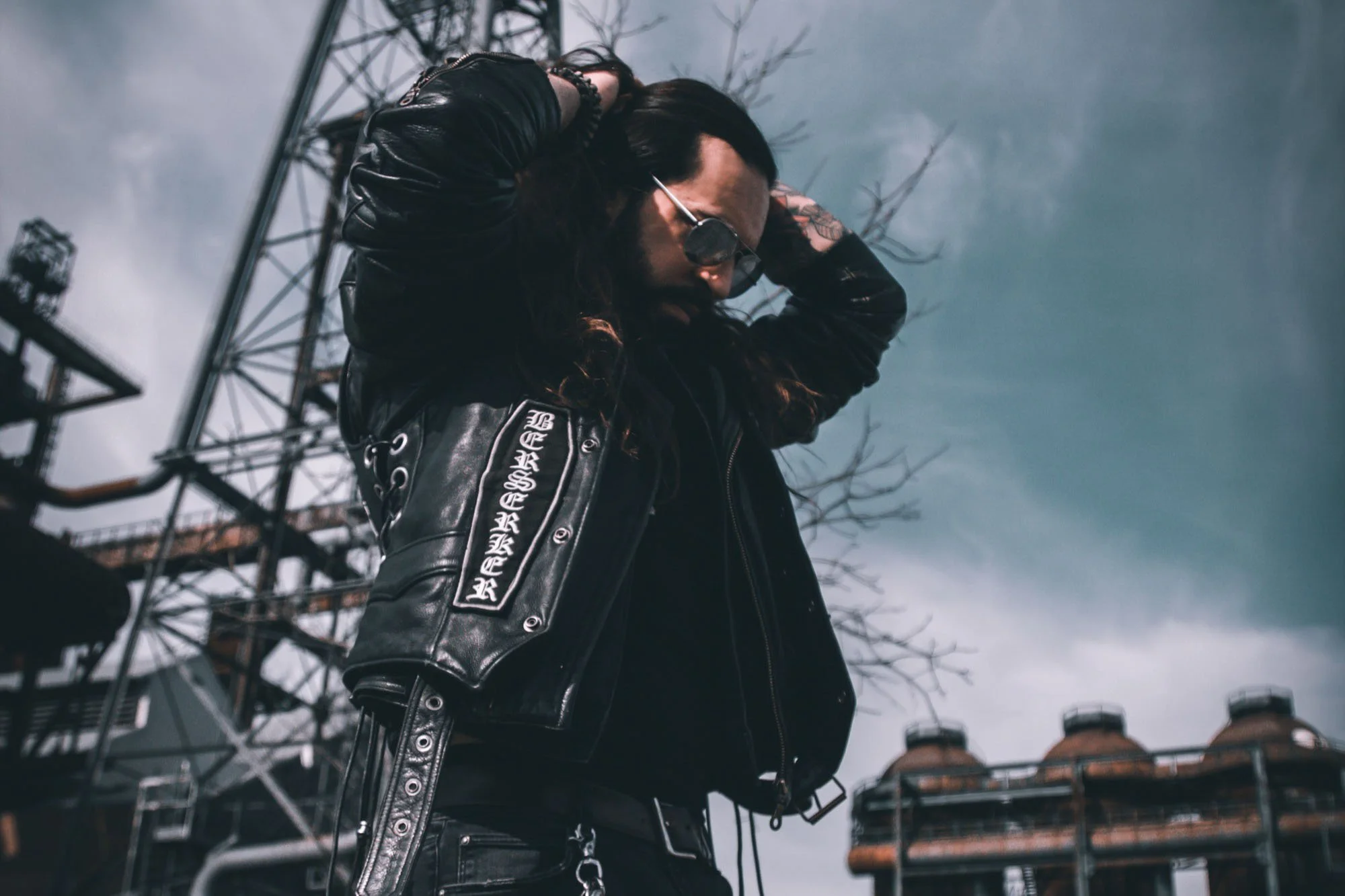 Junge Person in schwarzer Lederjacke mit Aufnähern, Sonnenbrille, lange dunkle Haare, steht vor einer metallischen Industrieanlage, während sie die Hände im Haar hat, bei bewölktem Himmel.