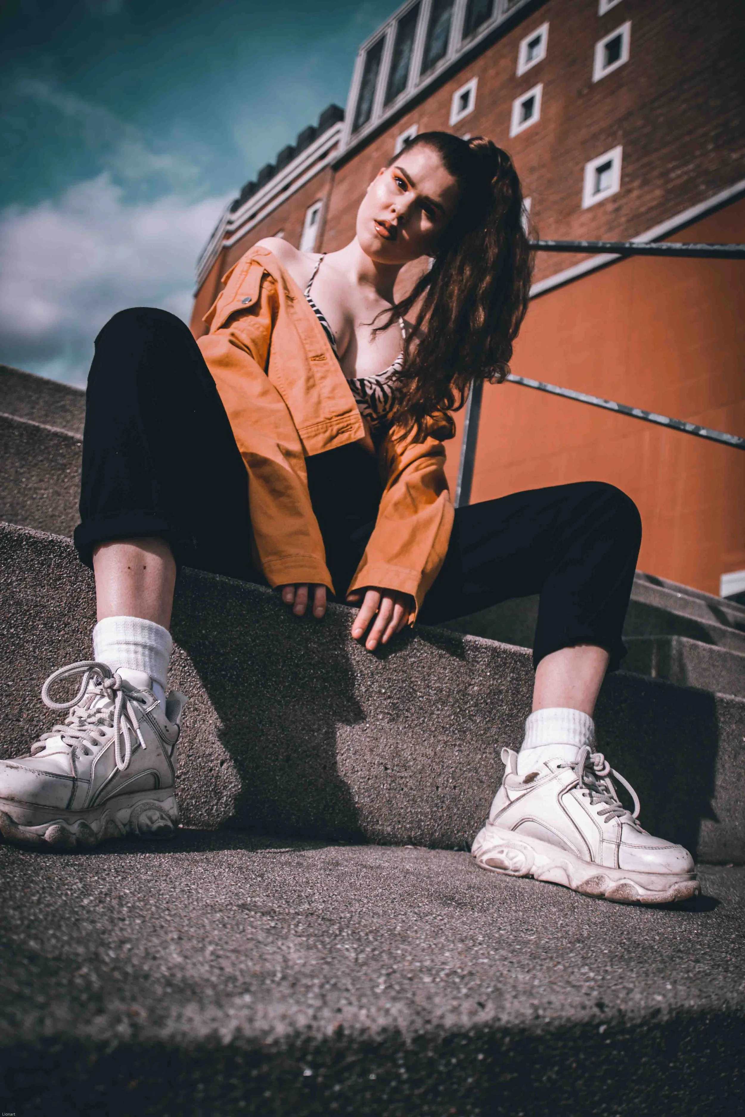 Junge Frau in Streetwear, sitzend auf Stufe vor einem modernen Gebäude, trägt weiße Turnschuhe, schwarze Hose, gelbe Jacke und hat lange braune Haare, bei Sonnenuntergang im urbanen Umfeld.