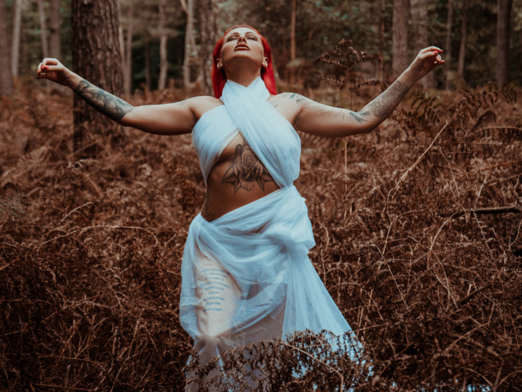 Frau mit roten Haaren in weißem Tuch, das um den Körper gewickelt ist, in einem Wald, Arme ausgestreckt, lebt.
