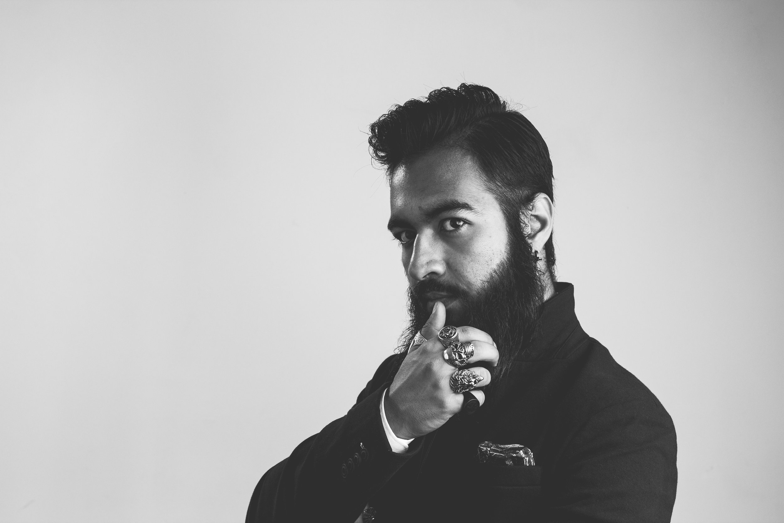 Schwarz-weiß Porträt eines Mannes mit stoppelndem Bart, der eine Hand mit mehreren Ringen an seinem Kinn hält und direkt schaut.