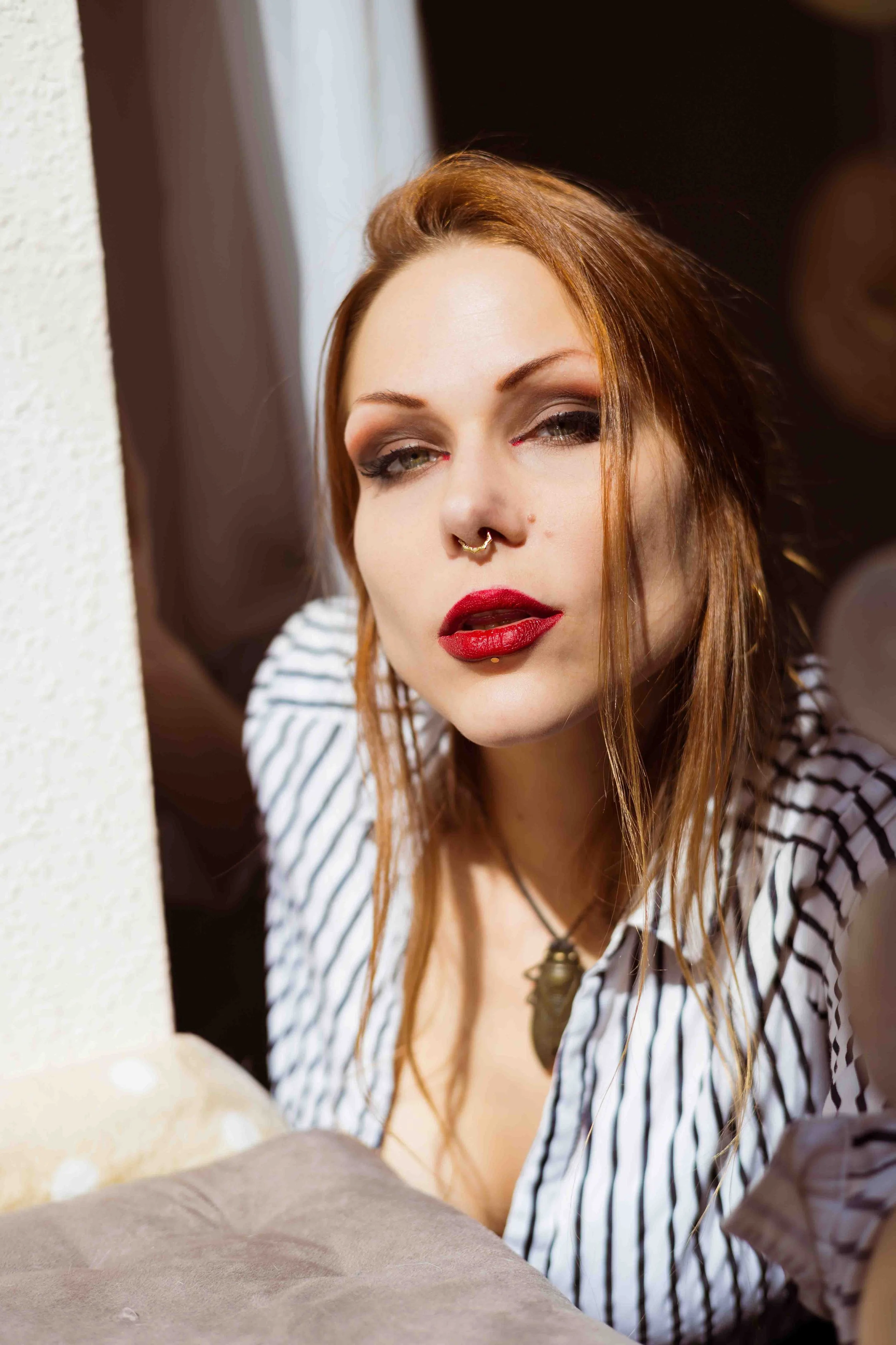 Nahaufnahme einer jungen Frau mit roten Haaren, Makeup und Piercings, trägt ein gestreiftes Hemd und eine Halskette, lehnt an einer Wand.