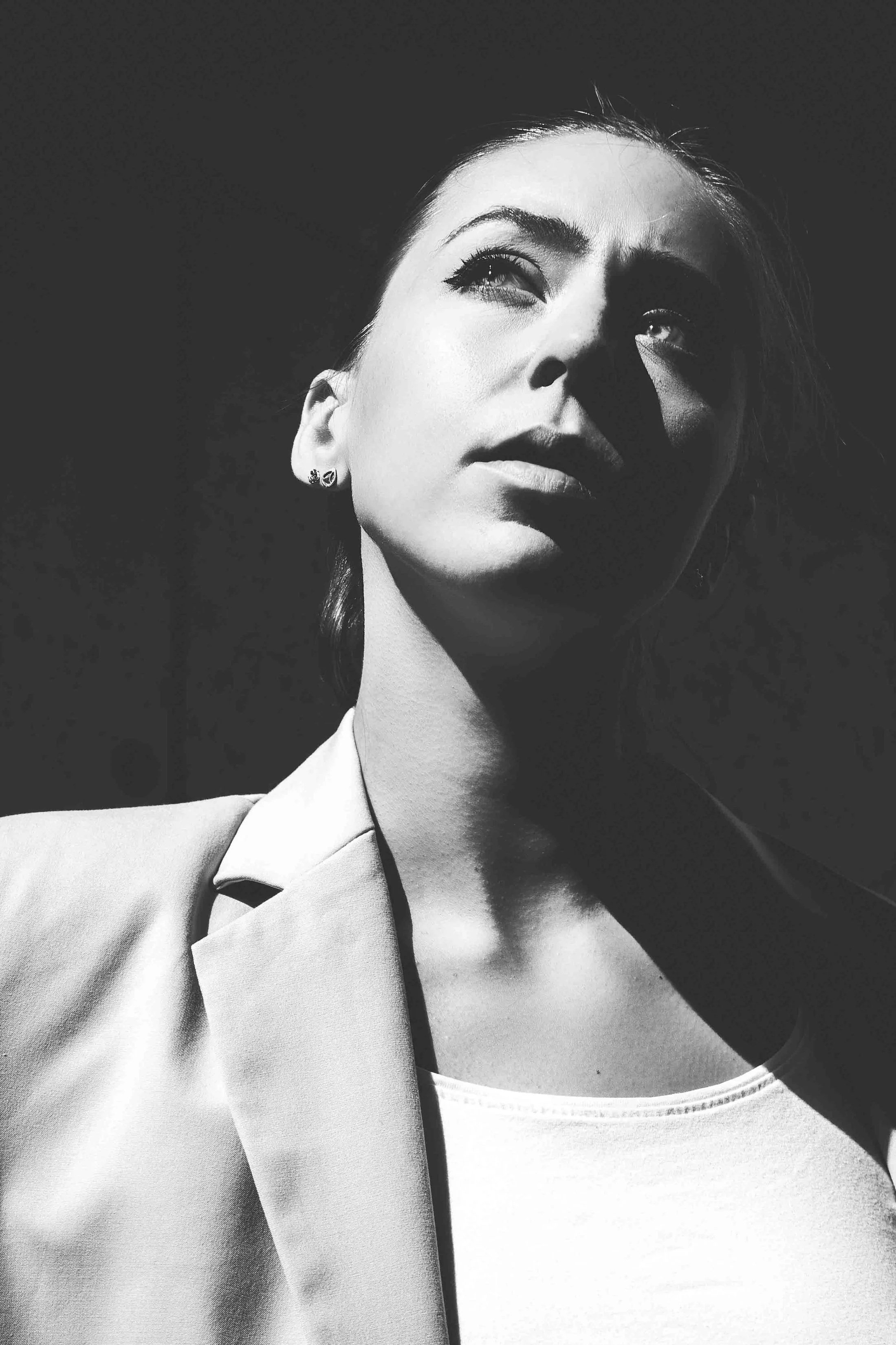 Schwarz-weiß Portrait einer jungen Frau mit kurz lockigem Haar, trägt eine Jacke und ein T-Shirt, blickt nach oben, Schatten betont ihr Gesicht.