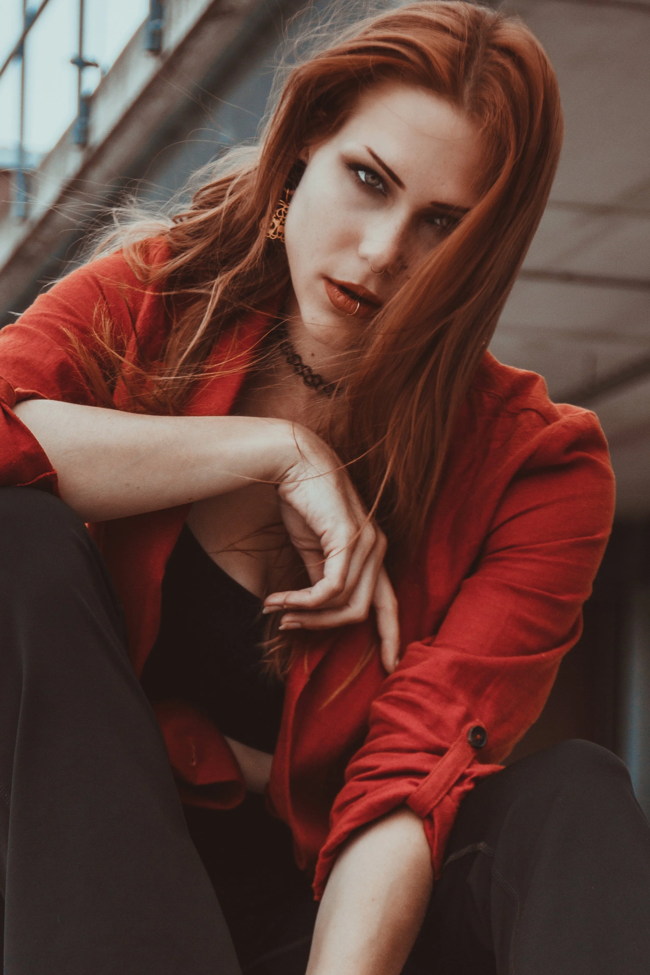 Junge Frau mit rotem Haar, dunklem Top und rotem Hemd, sitzend auf einer Treppe, schaut in die Kamera, im urbanen Umfeld.