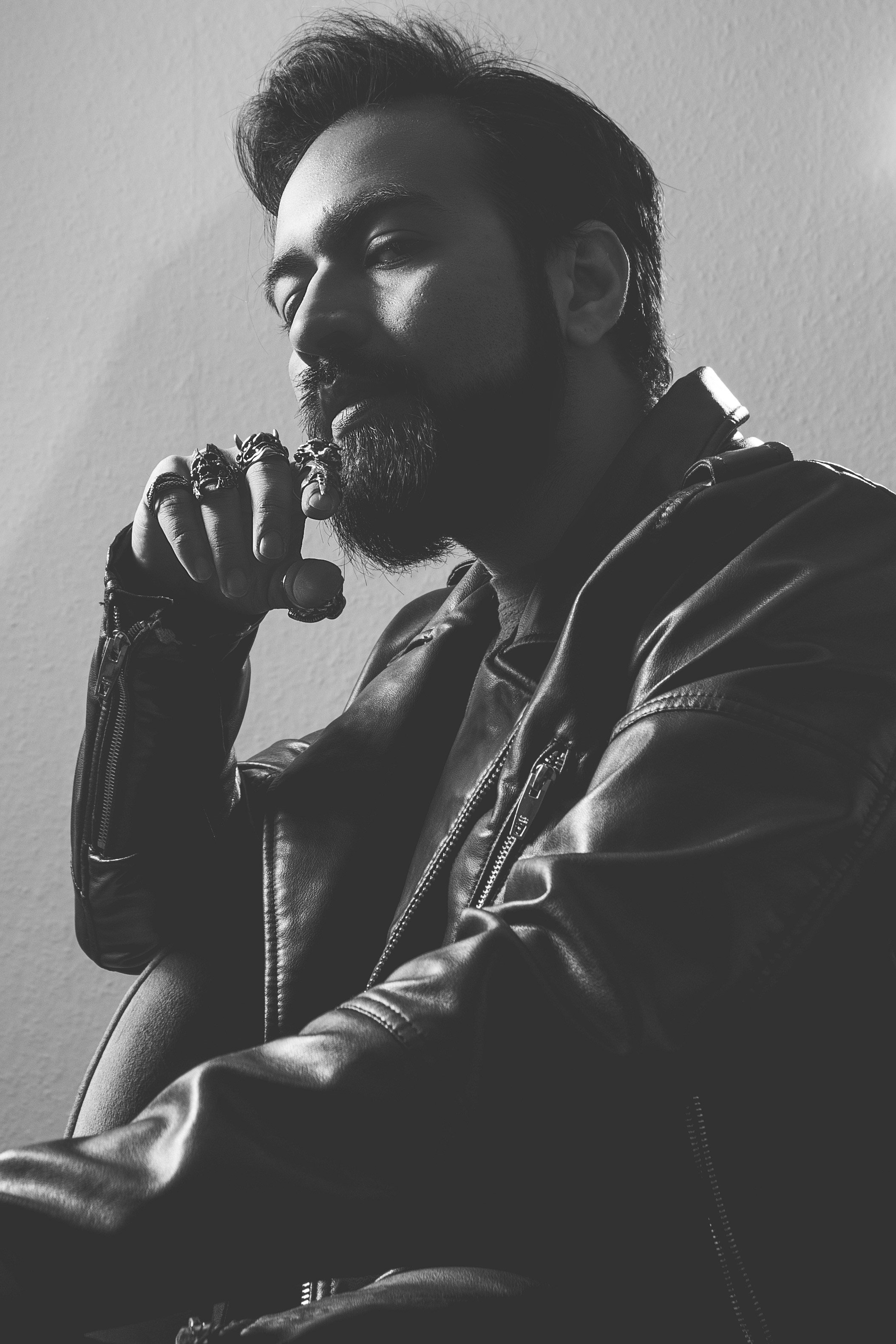 Schwarz-weißer Porträtfotografie eines Mannes mit Bart, der eine Lederjacke trägt und nachdenklich aussieht, während er eine Hand mit Ringen an seinem Kinn stützt.