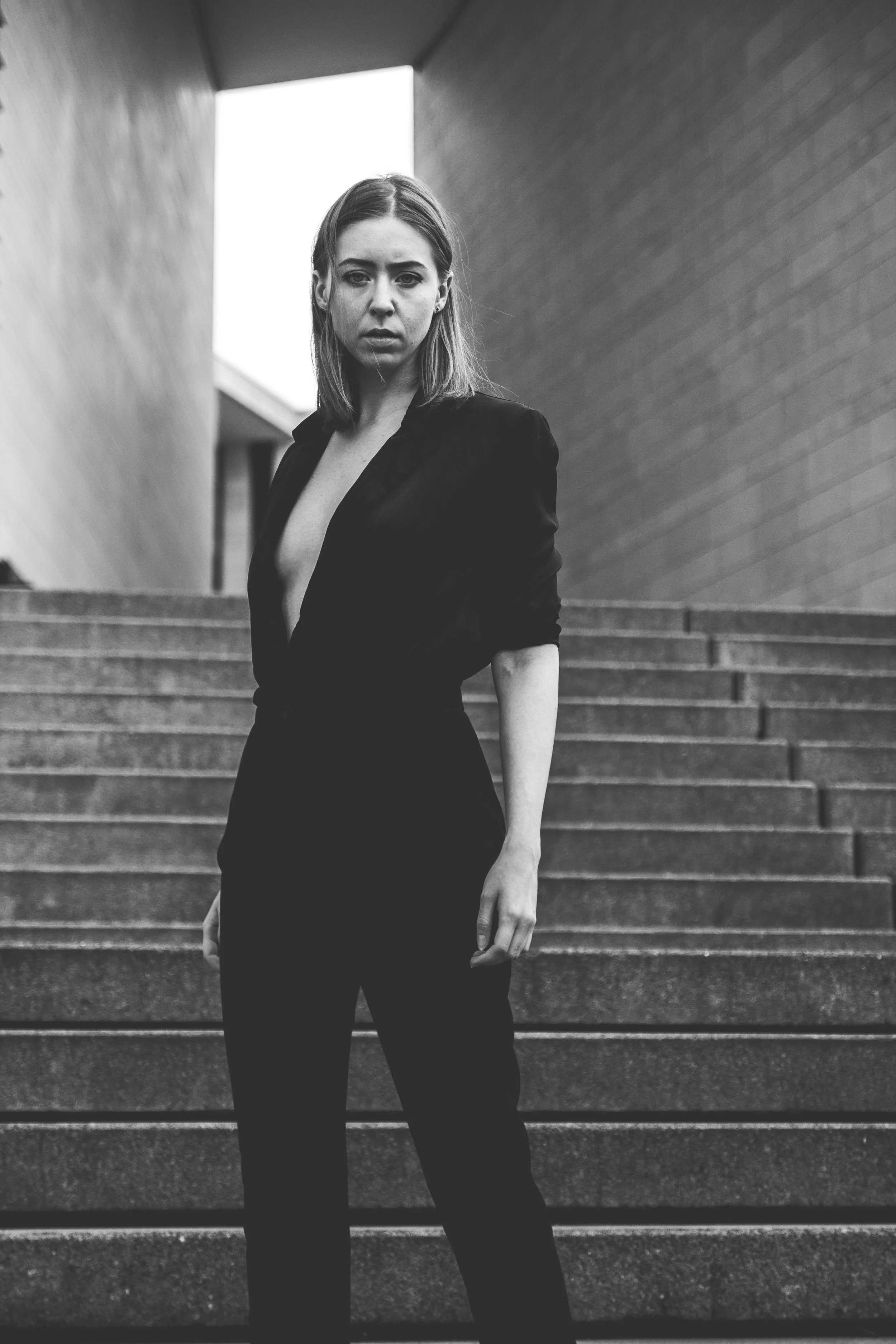 Schwarz-weiße Fotografie einer jungen Frau in schwarzem Anzug, stehend auf einer Treppe zwischen zwei modernen Gebäuden, mit ernstem Ausdruck, Blick in die Kamera.