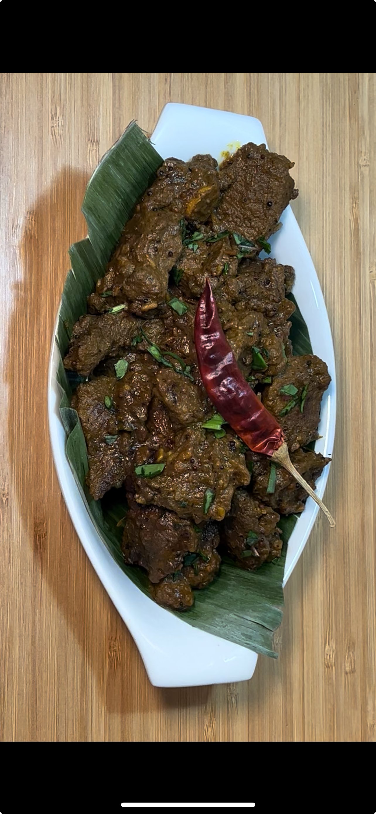 Plate of cooked beef curry garnished with chopped herbs and a dried red chili, served on a banana leaf-lined white dish on a wooden table.
