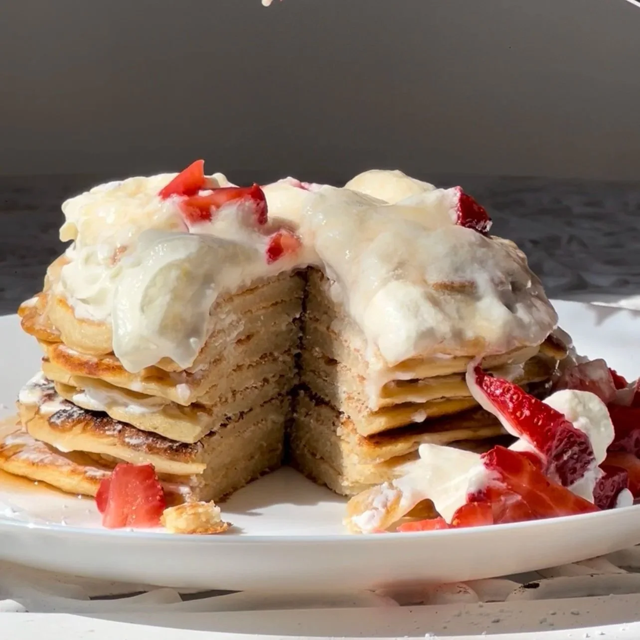 Stacked pancakes with strawberries, whipped cream, and syrup on a white plate near the water.