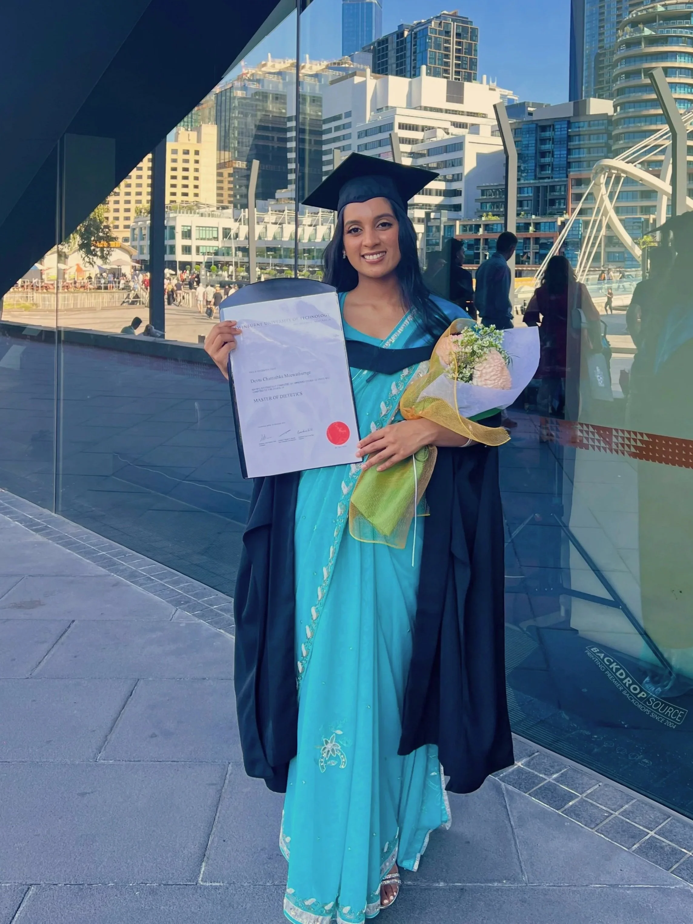 A young woman in graduation gown and cap holding a diploma and a bouquet of flowers outside in an urban area with tall buildings and glass architecture.
