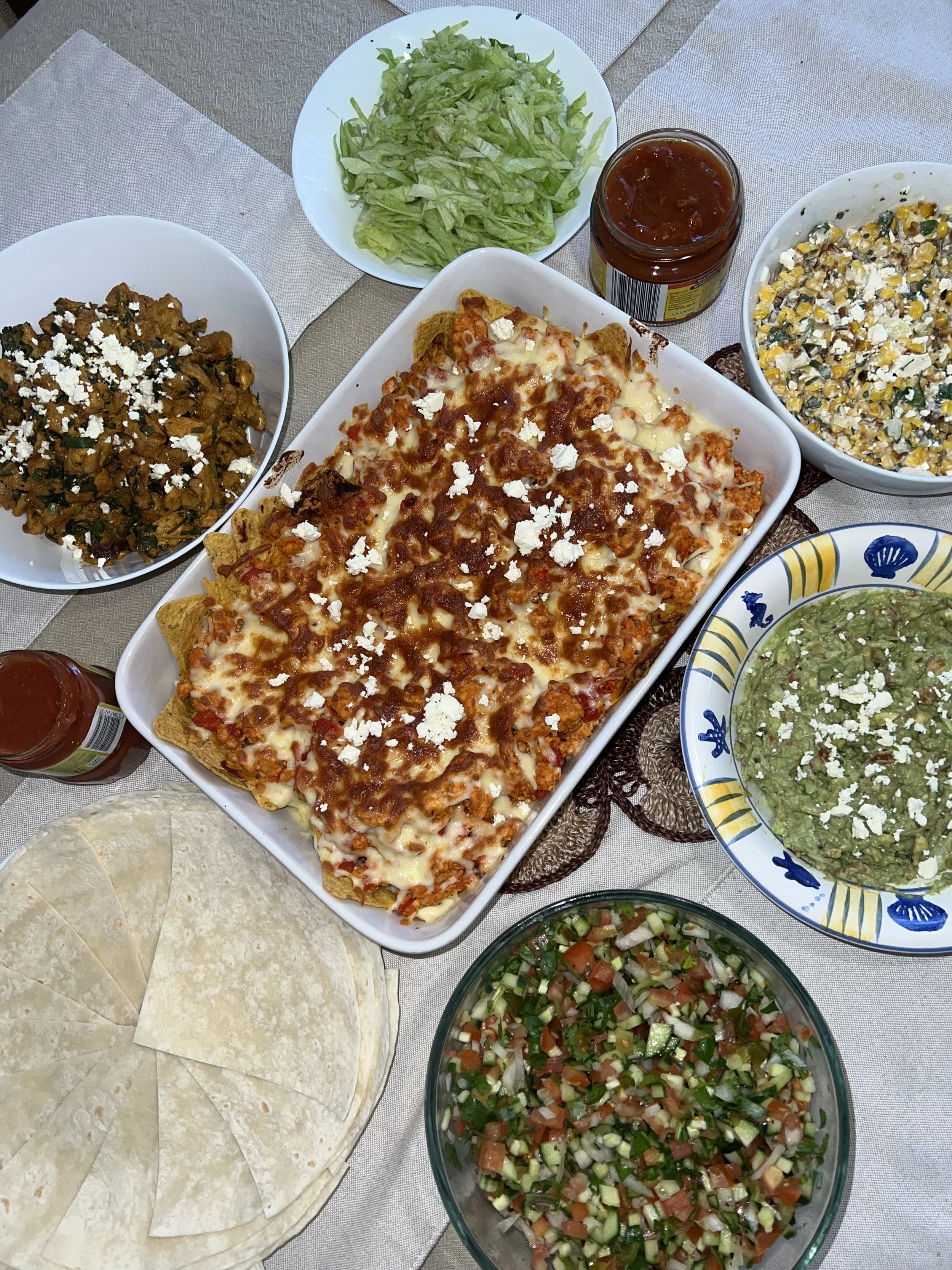 A table with various Mexican dishes, including a large baked dish of chicken casserole topped with cheese, bowls of chopped lettuce, corn salad, guacamole, pico de gallo, shredded chicken with vegetables, tortillas, and salsa.