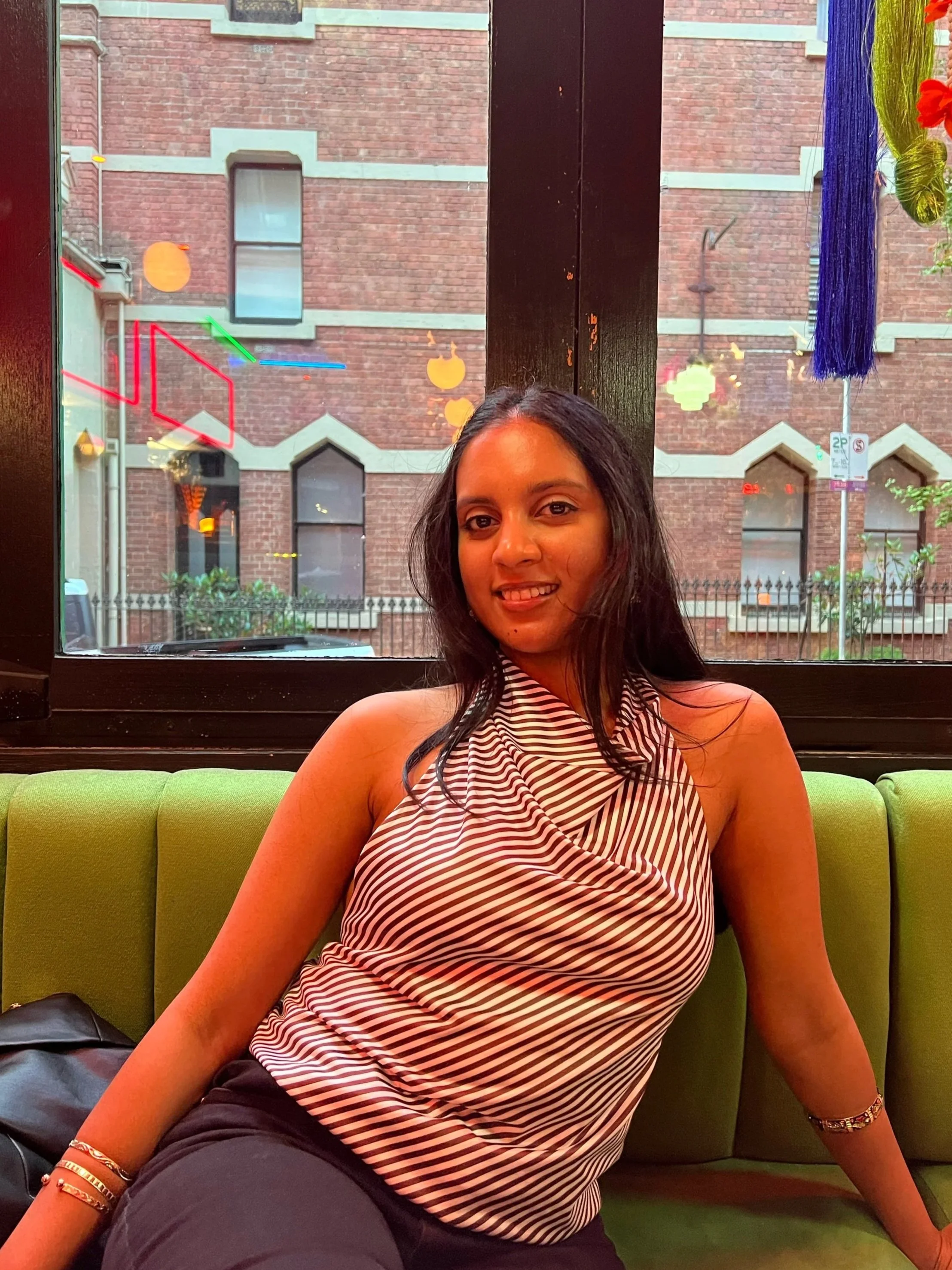 A woman with long dark hair sitting on a green booth seat in a restaurant, smiling, with a window behind her showing a brick building and colorful hanging decorations outside.