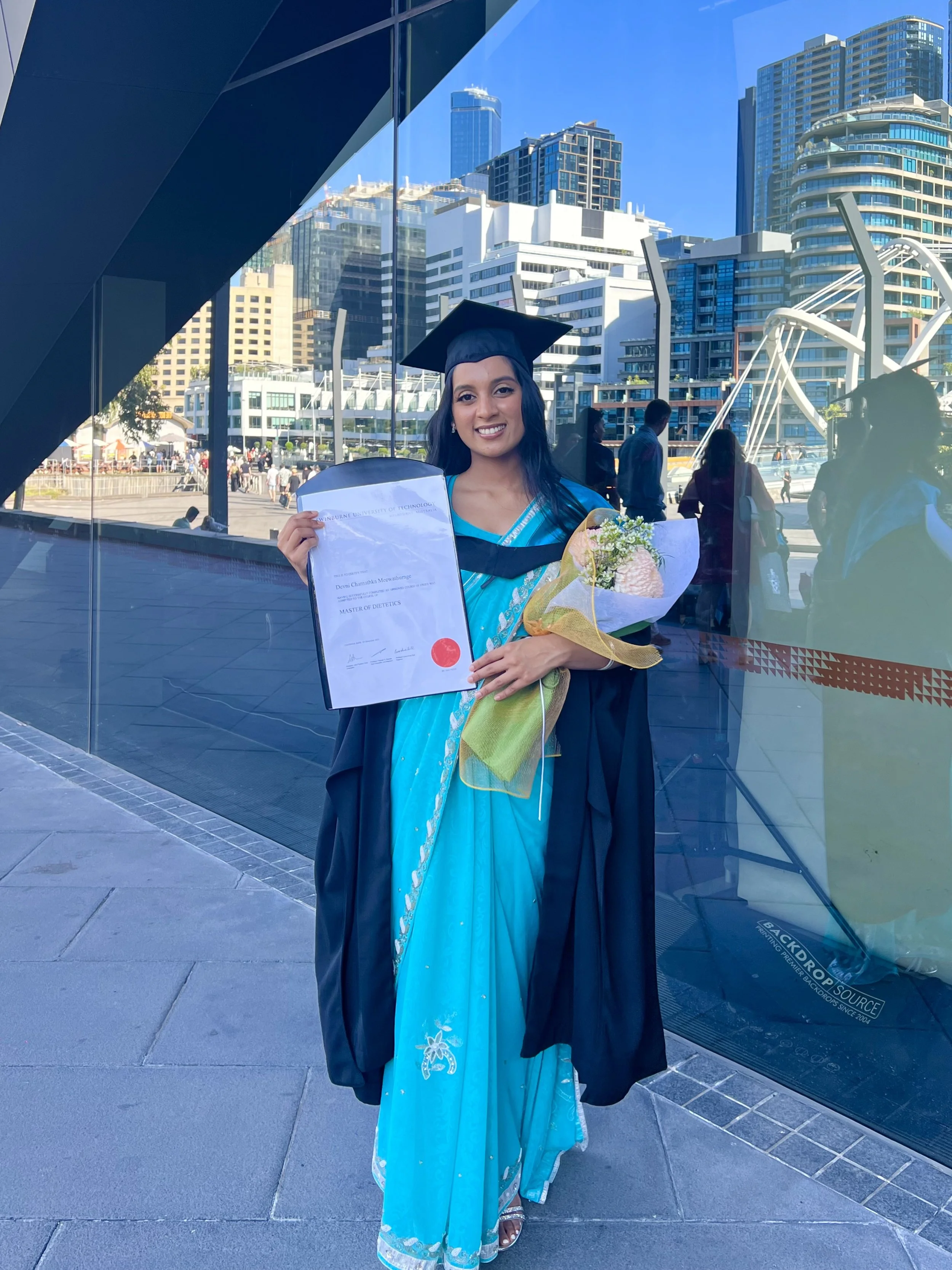 Woman in graduation cap and gown holding a diploma and bouquet of flowers, standing outdoors in a city with modern high-rise buildings reflected in glass.