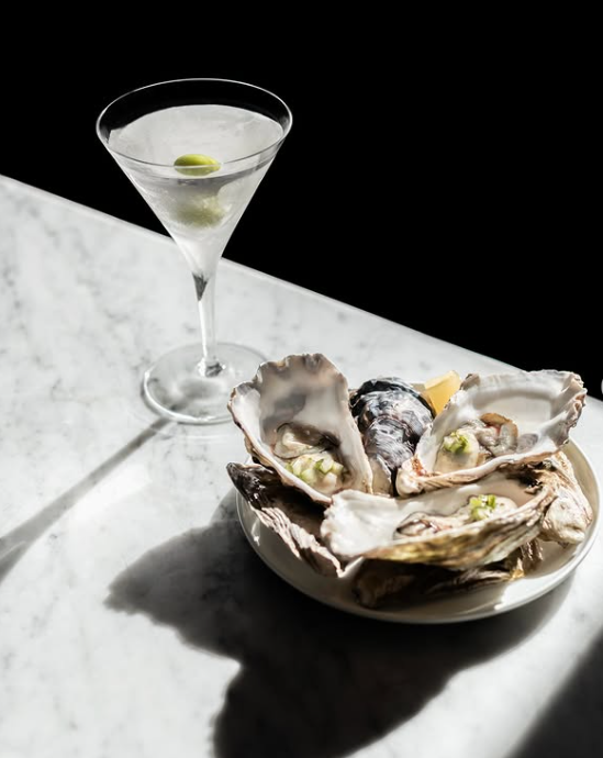 A glass of martini with green olives and a plate of oysters with lemon on a white marble surface.