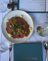 A plate of raw beef carpaccio topped with microgreens and capers, garnished with lemon wedges, set on a table with a glass of water, a blue notebook, and cutlery.
