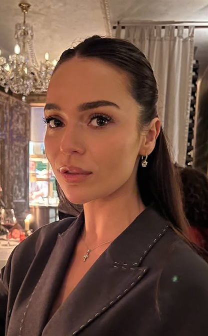 A woman with long dark hair tied back, wearing earrings and a dark blazer, posed indoors with a chandelier and curtains in the background.