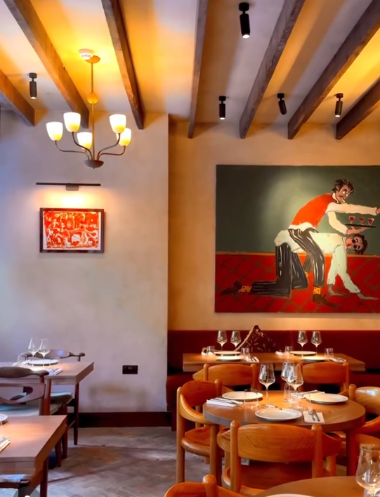 Interior of a restaurant with wooden ceiling beams, modern chandeliers, and artwork on the walls. The dining area has tables set with plates, wine glasses, and utensils, and the chairs are wooden with orange cushions.