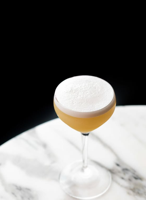 A cocktail in a stemmed glass with frothy white foam on top, placed on a white marble surface against a black background.