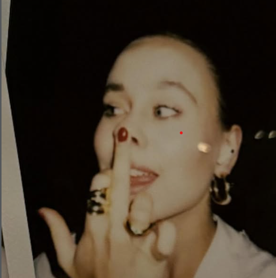 A woman with dark hair with a side part, wearing earrings, making a gesture by placing her index finger vertically in front of her lips and looking to her left. She has multiple rings on her fingers and is dressed in a white top.
