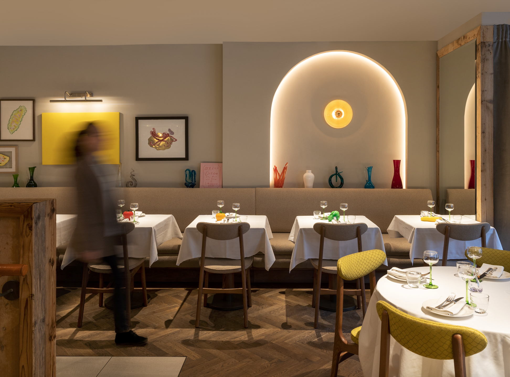 A restaurant dining area with tables set with white tablecloths, glassware, and napkins, surrounded by wooden chairs with yellow-patterned cushions. There is a beige cushioned banquette along the wall, decorated with colorful vases and framed artwork. A large mirror and a lit arched wall feature with a circular light fixture are visible in the background. A person moving past the camera appears blurred.