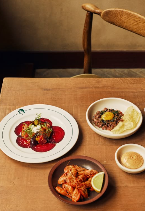 Three dishes of food on a wooden table: a plate of beet salad with greens and garnish, a bowl of tartare with chips and a dab of sauce, and a small bowl of dipping sauce with a lime wedge.