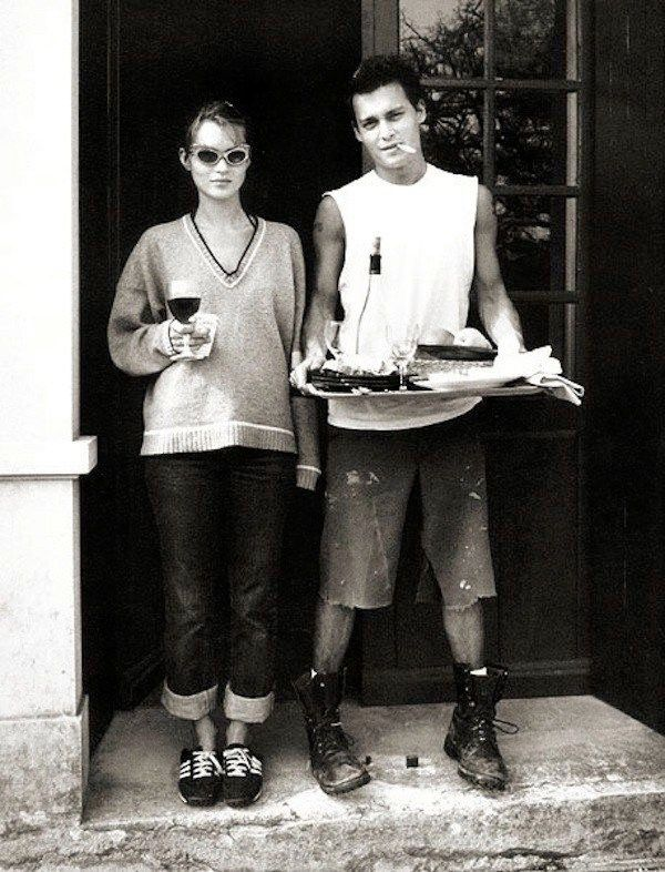 A young woman with sunglasses holding a glass of wine, standing next to a young man with dark hair, wearing ripped clothes and boots, holding a tray with food or objects, outside a building.