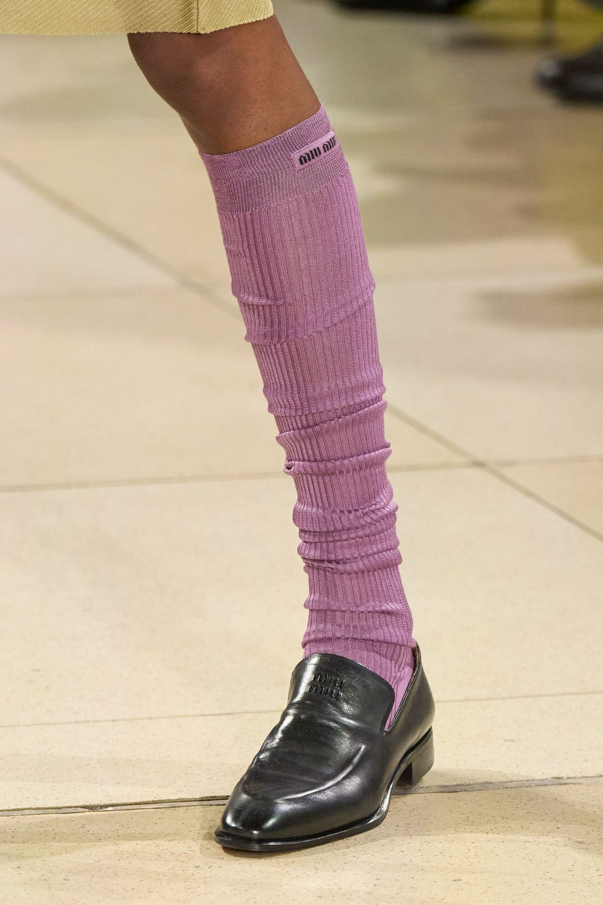 Close-up of a person's leg wearing a purple sock with black text near the top, black shiny oxford shoes, standing on tiled floor.