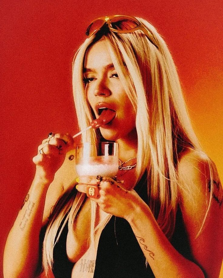 A woman with long blonde hair, wearing sunglasses on her head, wearing rings and a necklace, holding a cocktail glass with a drink, licking a lollipop, against a gradient orange-red background.