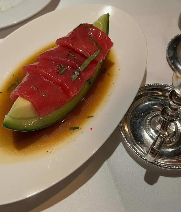 A plate with a halved cantaloupe filled with sliced prosciutto and garnished with herbs, served with a sauce, next to a silver candle holder.